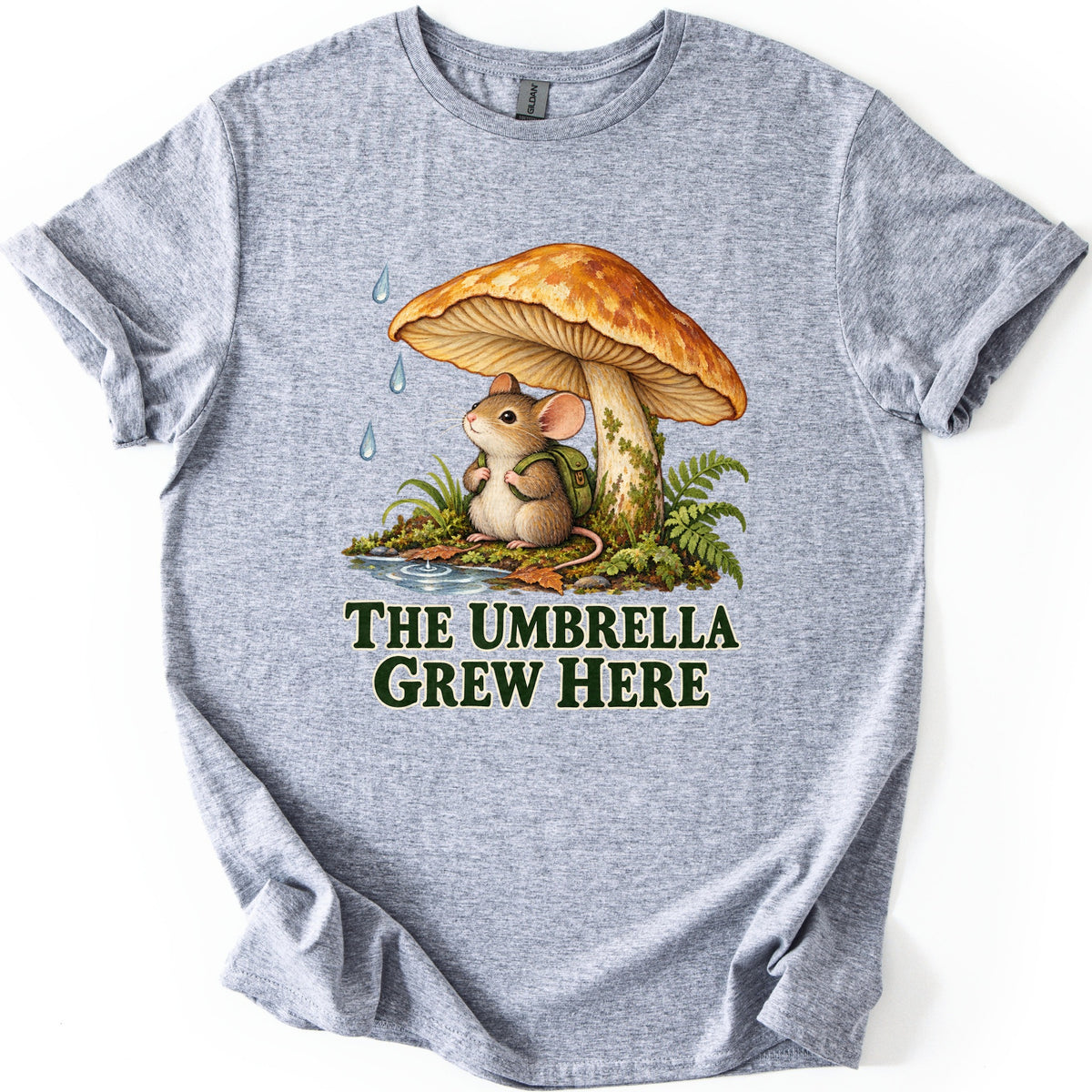 The Umbrella Grew Here Comfort Colors T-shirt in Sport Grey, laid flat. A mouse wearing a green backpack sits under a large orange mushroom as raindrops fall, beside ferns and a puddle, above the words The Umbrella Grew Here in dark green letters. Shown on a white background.