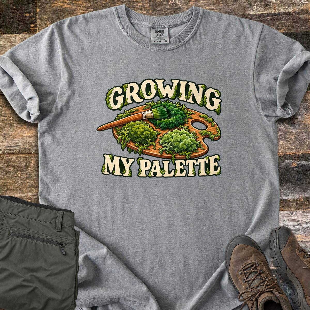 Growing My Palette Comfort Colors T-shirt in Grey, laid flat. A wooden painter's palette covered in green moss, with a brush whose bristles are moss, between the words Growing My Palette in cream letters. Olive pants and brown hiking boots lie beside it on weathered wood.