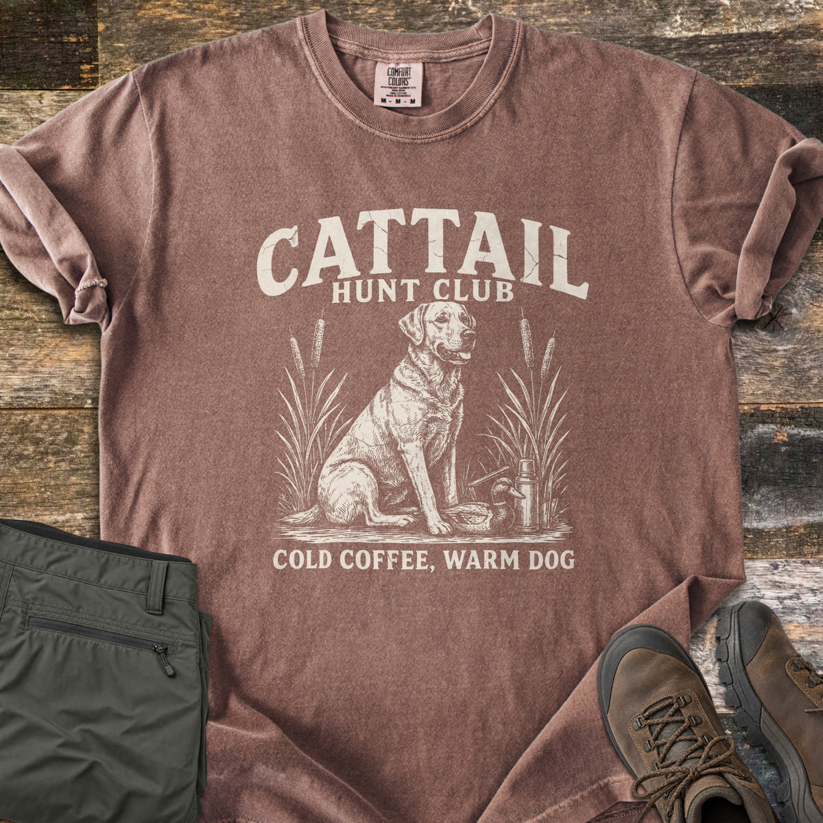 Cattail Hunt Club Comfort Colors T-shirt in Espresso, laid flat. Cream print of a Labrador sitting among cattails beside a duck decoy and thermos, under the words Cattail Hunt Club, above Cold Coffee, Warm Dog. On weathered wood with olive pants and brown hiking boots.