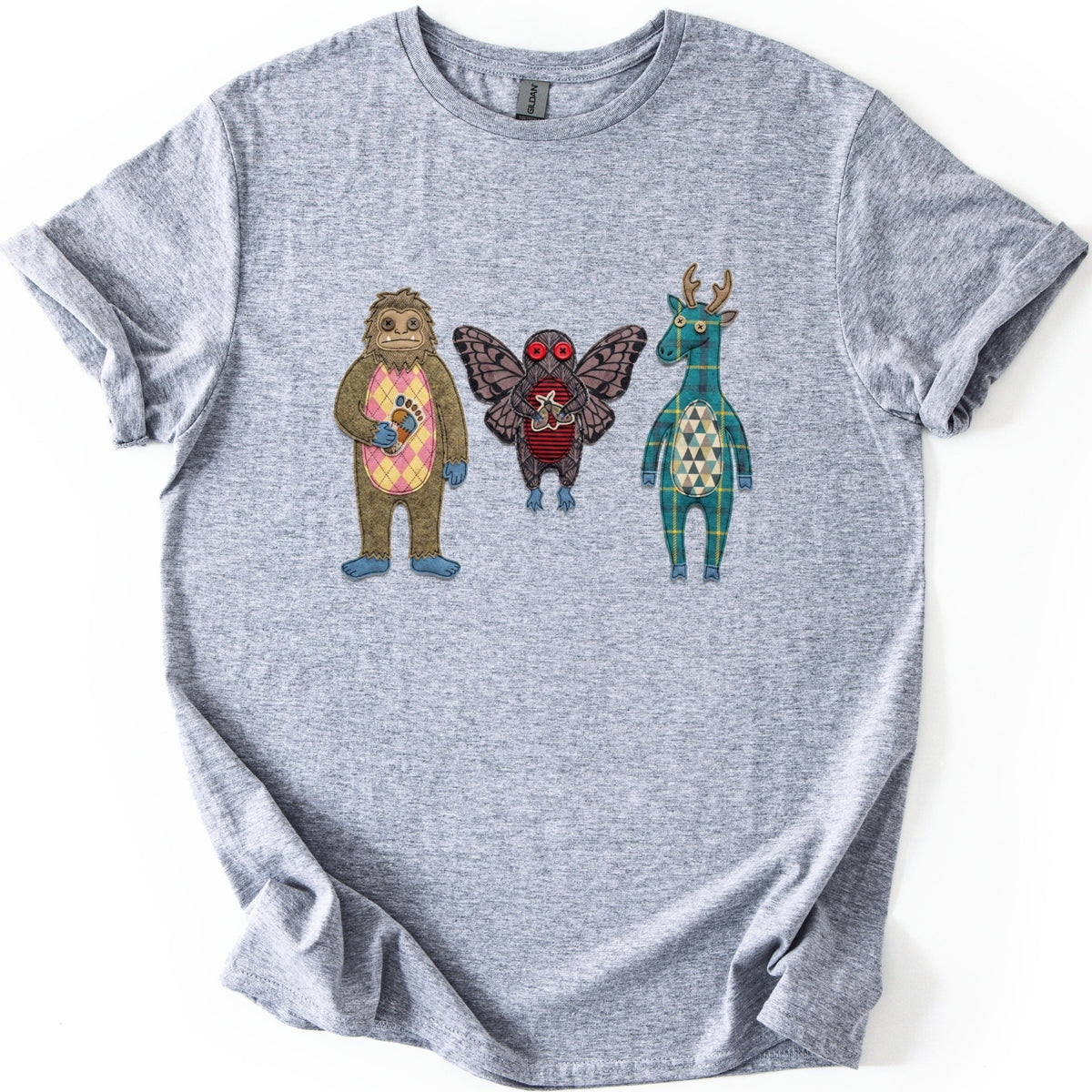 Patchwork Cryptid Trio Comfort Colors T-shirt in Sport Grey, laid flat. Three stitched patchwork cryptids with no text: a brown Bigfoot with a pink argyle belly, a winged Mothman with red button eyes, and a teal plaid antlered creature. Laid on a white surface.