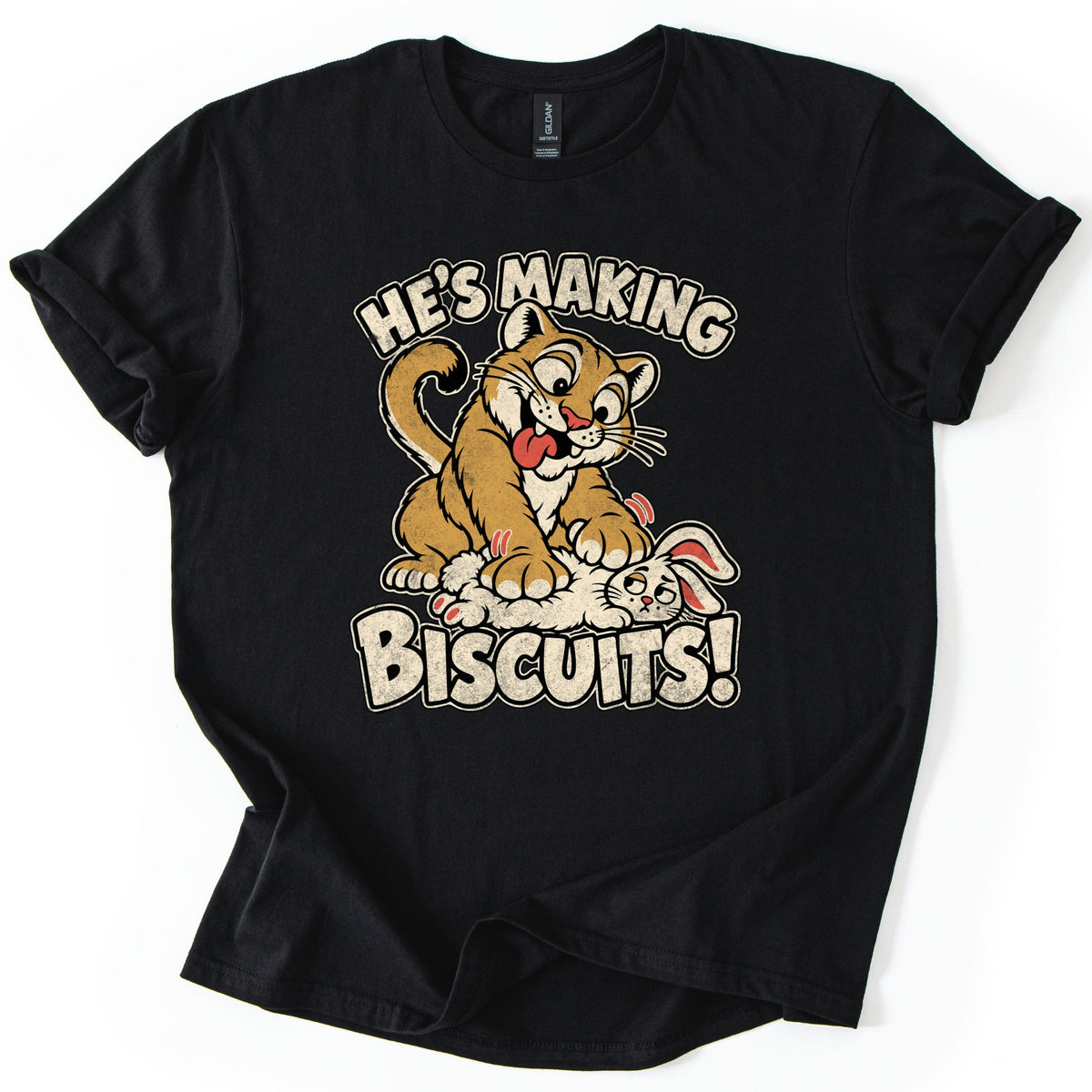 He's Making Biscuits Comfort Colors T-shirt in Black, laid flat. A grinning cartoon cougar with its tongue out kneads a dazed white rabbit on its back, with the words He's Making Biscuits! in distressed cream letters. The shirt lies on a white background with rolled sleeves.