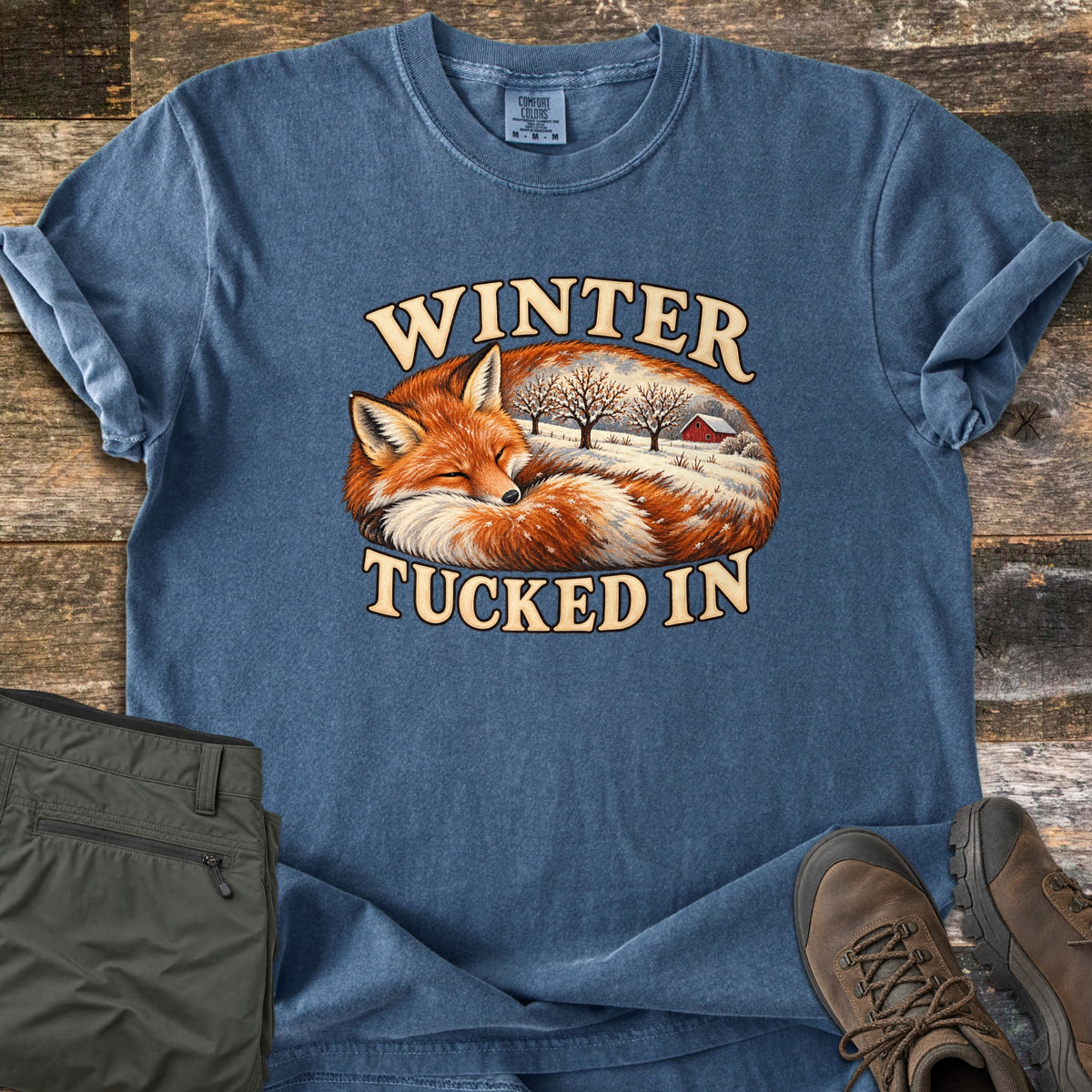 Winter Tucked In Comfort Colors T-shirt in Denim, laid flat. A painted red fox sleeps curled up, its body filled with a snowy farm scene with bare trees and a red barn, between the words Winter Tucked In. Olive pants and brown hiking boots lie beside it on weathered wood.