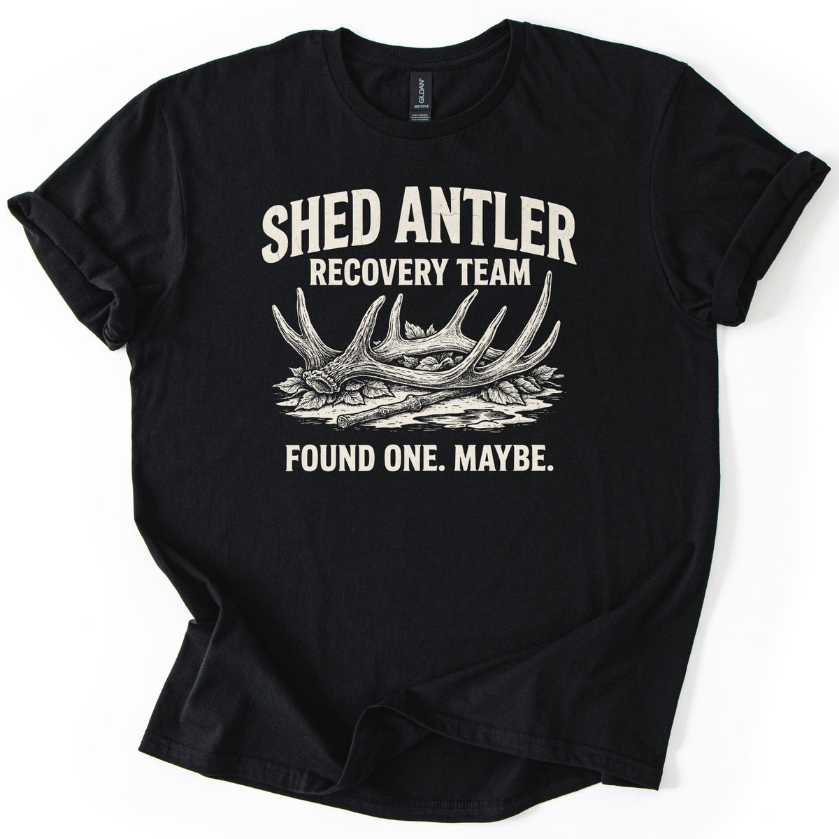 Shed Antler Recovery Team Comfort Colors T-shirt in Black, laid flat. Cream text reads Shed Antler Recovery Team above a detailed sketch of a fallen deer antler resting on leaves beside a twig, with Found One. Maybe. below. The sleeves are rolled on a white surface.