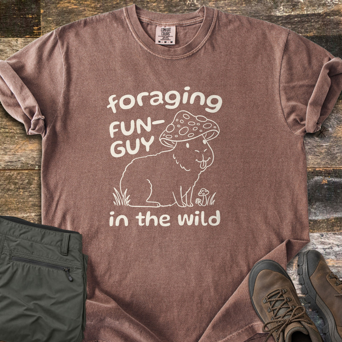 Foraging Fun-Guy in the Wild Comfort Colors T-shirt in Espresso, laid flat. A cream line drawing of a capybara wearing a spotted mushroom cap, tongue out, sits in grass by tiny mushrooms between the words foraging FUN-GUY in the wild. Olive hiking pants and brown boots lie on weathered wood.