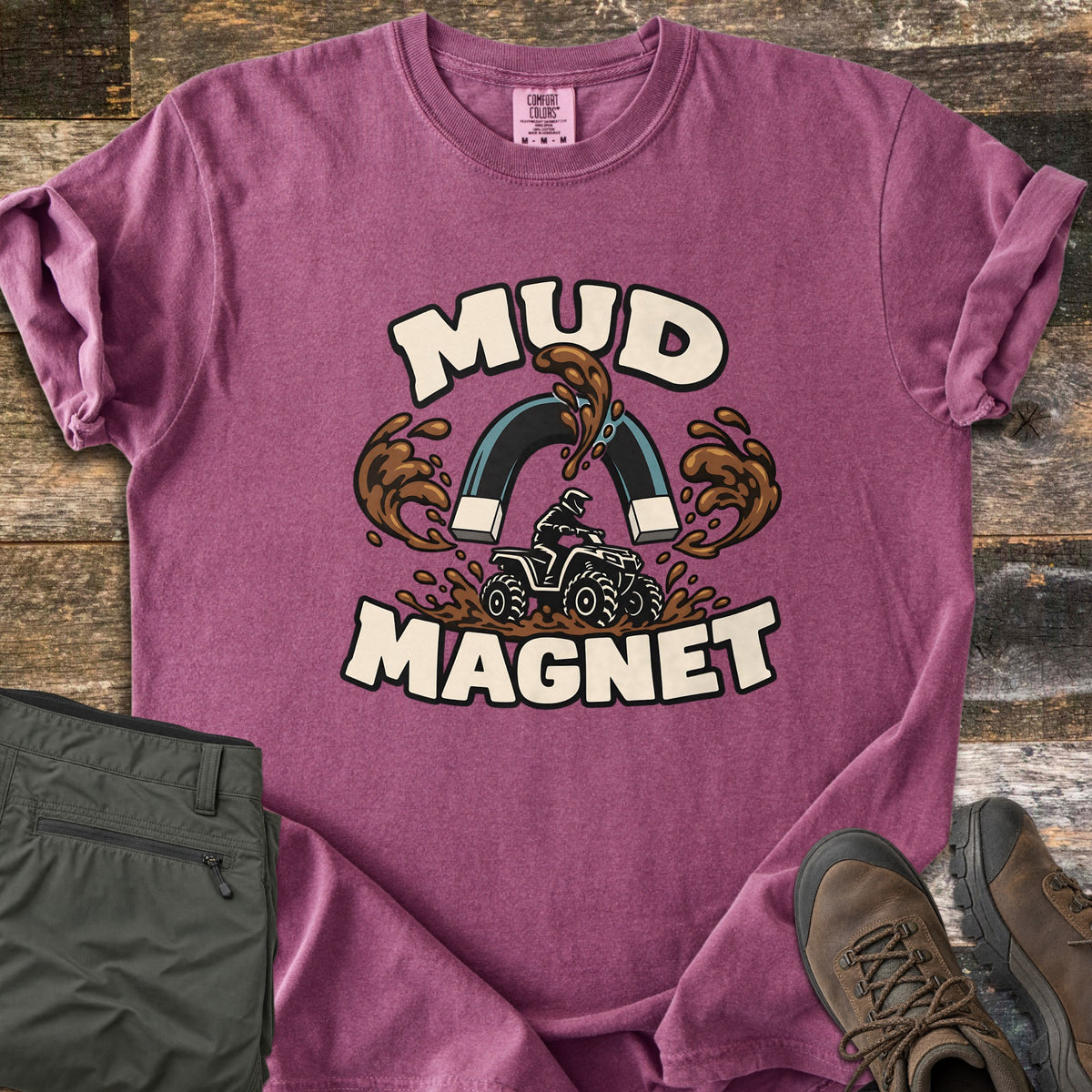 Mud Magnet Comfort Colors T-shirt in Berry, laid flat. The words MUD MAGNET arch around a horseshoe magnet pulling mud splashes toward an ATV rider churning through mud. Shown on a wood floor with folded olive pants and brown hiking boots.