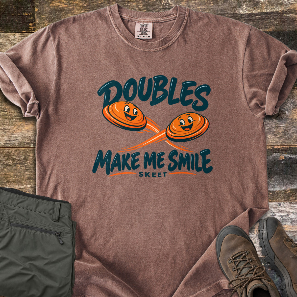 Doubles Make Me Smile Comfort Colors T-shirt in Espresso, laid flat. Two smiling orange clay targets fly on crossing paths between the words Doubles Make Me Smile, with Skeet printed below. Folded olive pants and brown hiking boots sit beside it on weathered wood planks.
