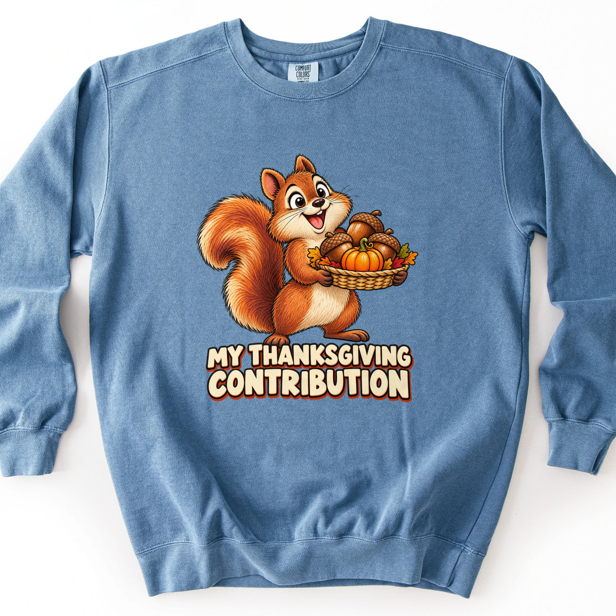 My Thanksgiving Contribution Comfort Colors crewneck sweatshirt in Blue Jean, laid flat. A smiling cartoon squirrel holds a woven basket of acorns, a small pumpkin, and autumn leaves above the words My Thanksgiving Contribution in cream letters. Shown on a plain white background.