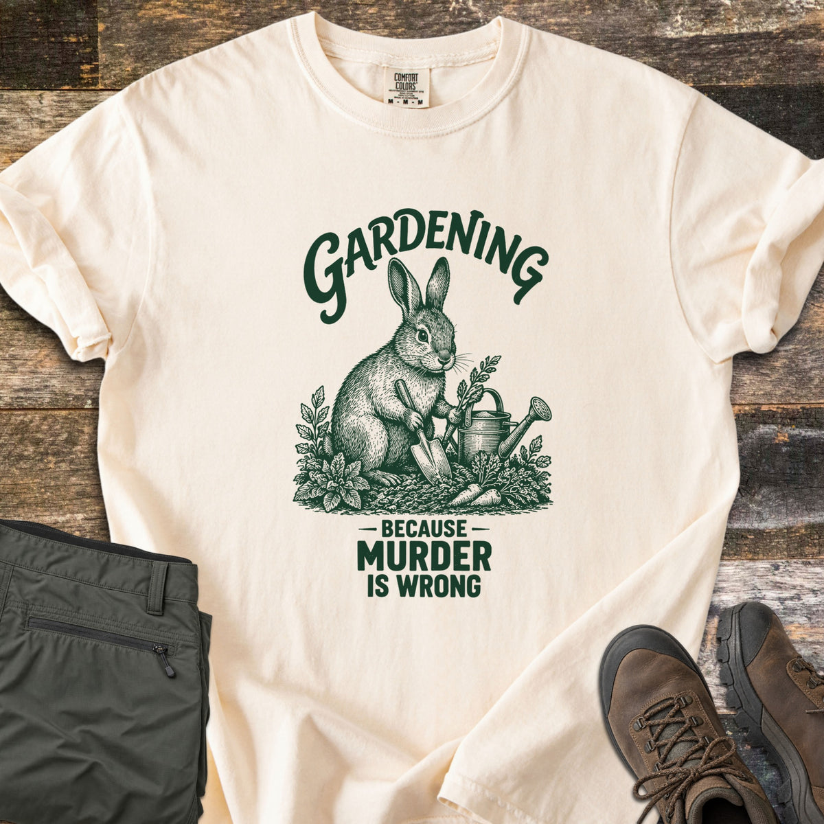 Gardening Murder Rabbit Comfort Colors T-shirt in Ivory, laid flat. A dark green etched-style rabbit holds a trowel beside a watering can, carrots and plants, under the word Gardening and above Because Murder Is Wrong. Green pants and brown hiking boots sit on weathered wood.