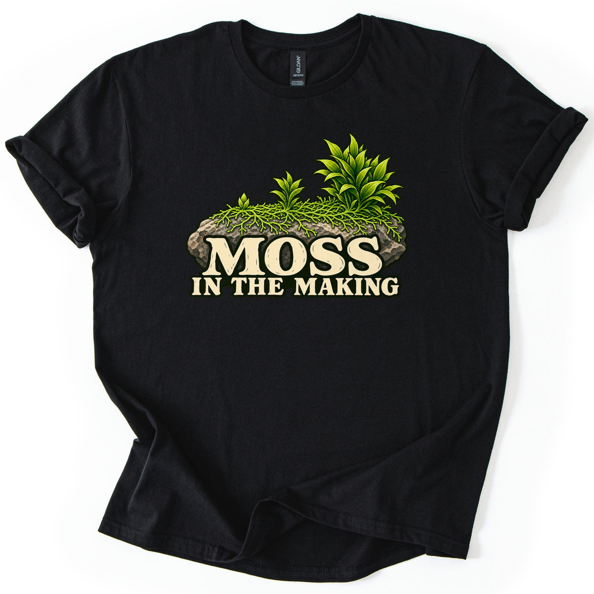 Moss in the Making Comfort Colors T-shirt in Black, laid flat. A mossy gray rock topped with leafy green plants sits above the words MOSS IN THE MAKING in cream lettering. The sleeves are rolled and the shirt lies on a white background.