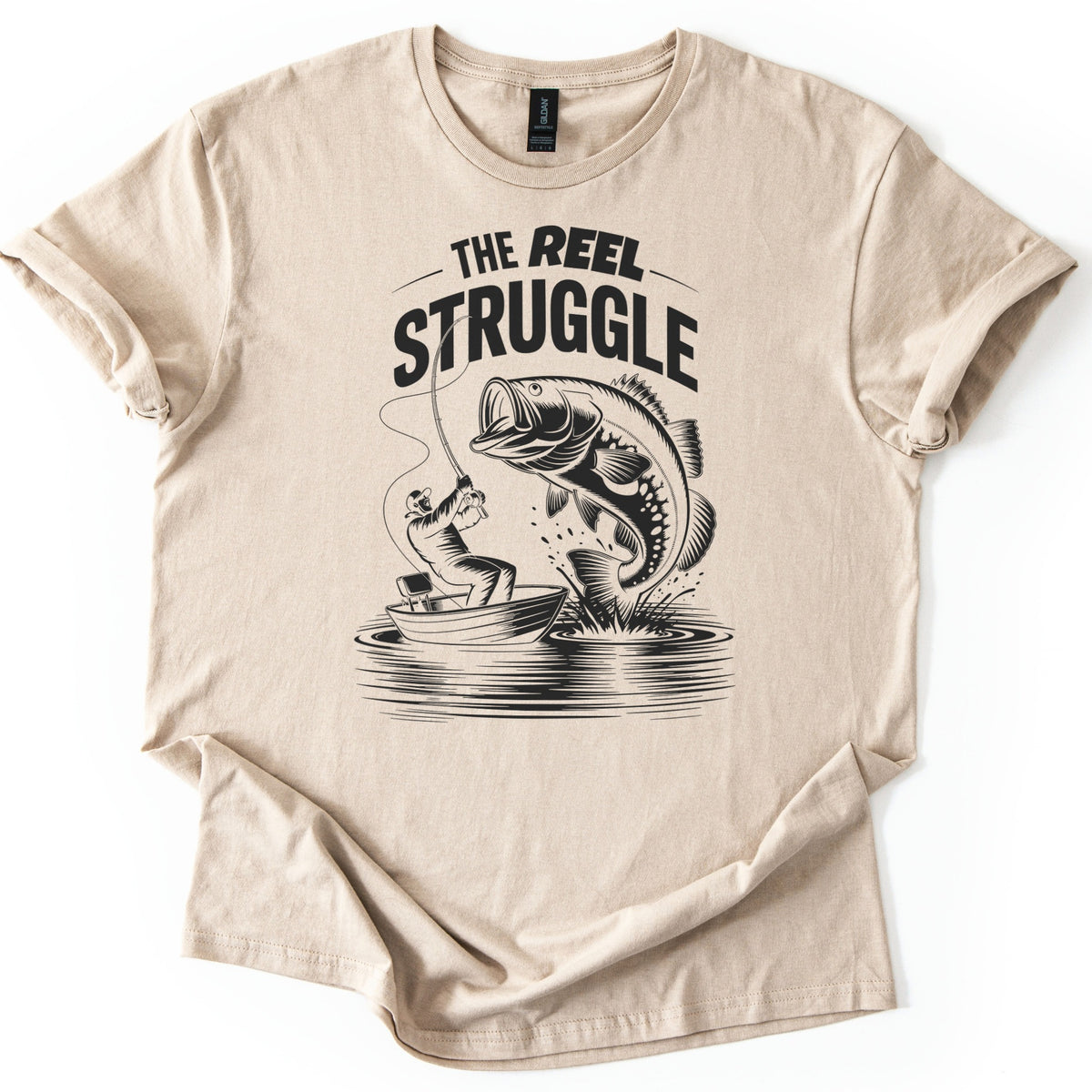 Reel Struggle Comfort Colors T-shirt in Sand, laid flat. Black print of the words The Reel Struggle above a fisherman in a small boat pulling back on a bent rod while a large bass leaps from the water. The sleeves are rolled, on a white background.