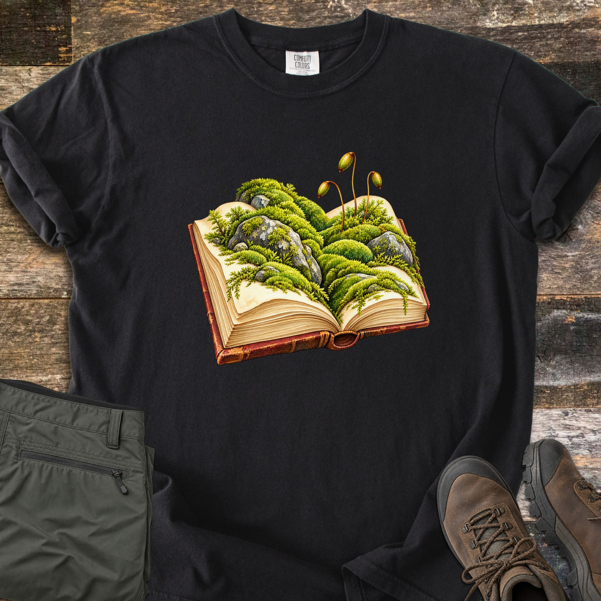 Moss Storybook Comfort Colors T-shirt in Black, laid flat. An illustrated open book with green moss, gray rocks, and tall moss sporophytes growing from its pages. Rolled sleeves, on weathered wood with folded olive pants and brown hiking boots.