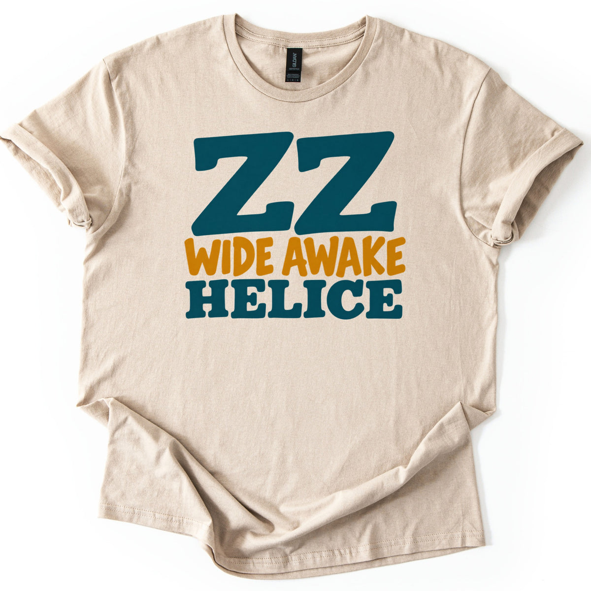ZZ Wide Awake Comfort Colors T-shirt in Sand, laid flat. Large dark teal letters ZZ sit above WIDE AWAKE in mustard yellow and HELICE in bold dark teal. The shirt lies on a white background with its sleeves cuffed and the hem folded in soft wrinkles.