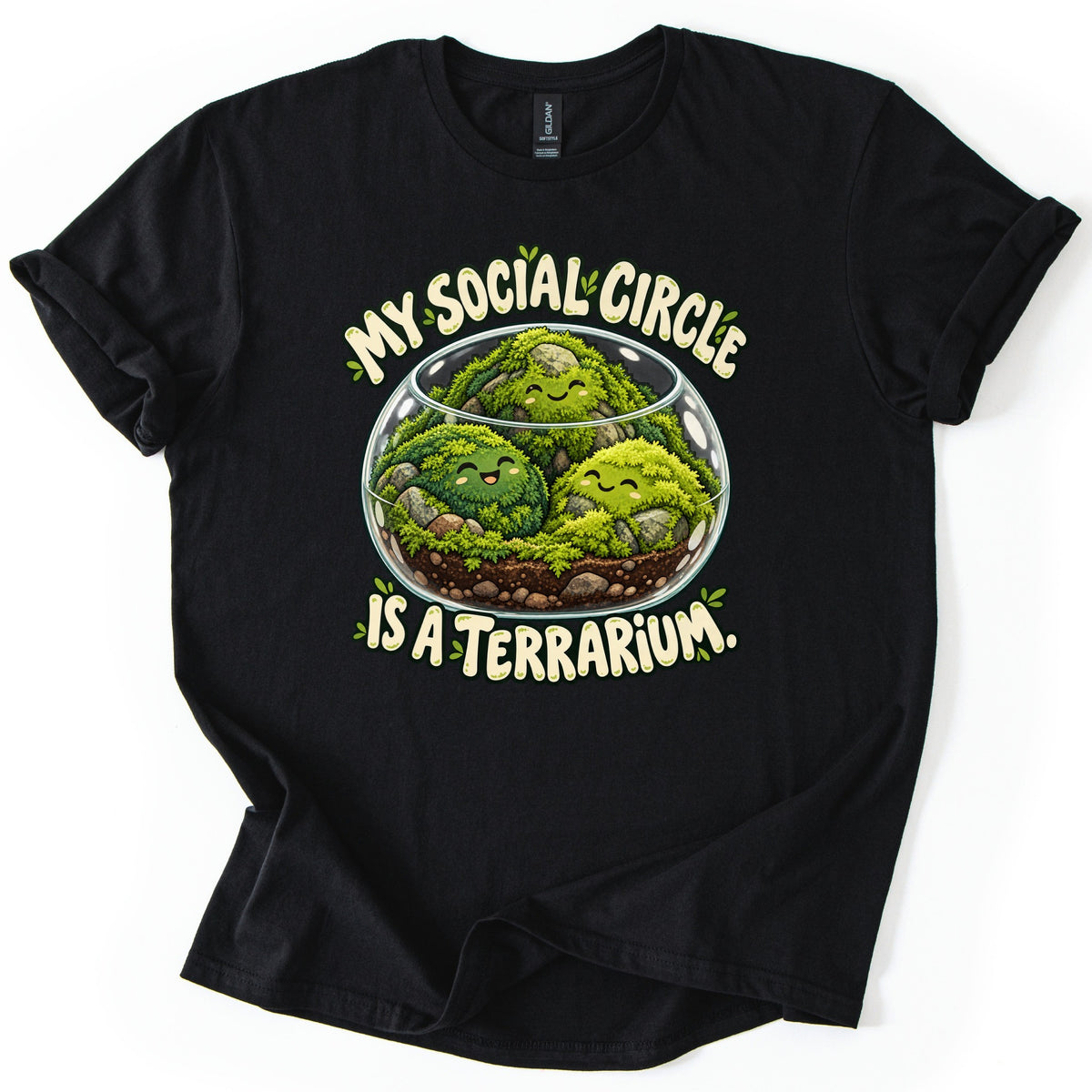 My Social Circle Is a Terrarium Comfort Colors T-shirt in Black, laid flat. A round glass terrarium holds three smiling green moss balls on soil and pebbles, framed by the cream words My Social Circle above and Is a Terrarium. below. The shirt lies on a white surface with cuffed sleeves.