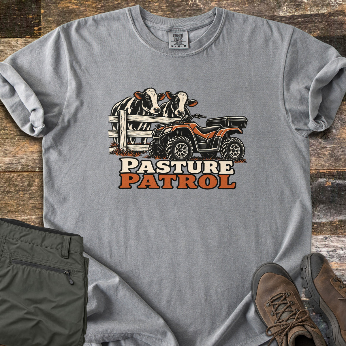 Pasture Patrol Comfort Colors T-shirt in Grey, laid flat. Two black and white cows lean over a wooden fence beside an orange and black ATV, above the words Pasture Patrol. Folded olive pants and brown hiking boots sit on a weathered wood surface.