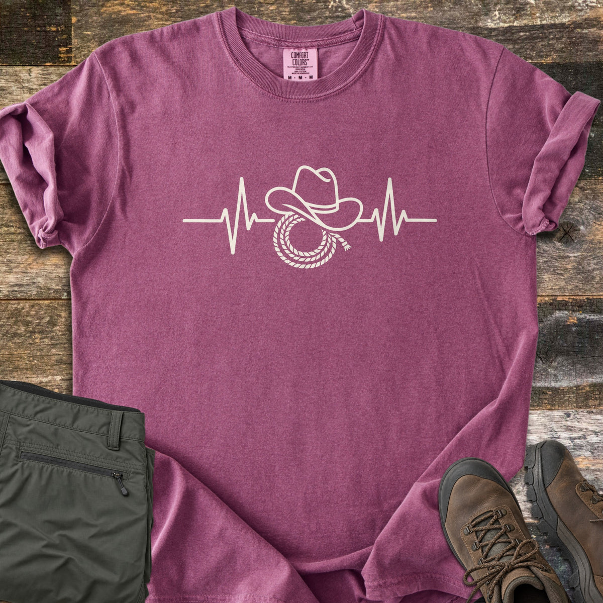 Cowboy Heartbeat Comfort Colors T-shirt in Berry, laid flat. A cream line art heartbeat line rises into peaks around a cowboy hat resting on a coiled lasso; there are no words. Sleeves are cuffed, on rustic wood boards beside folded olive cargo pants and brown hiking boots.