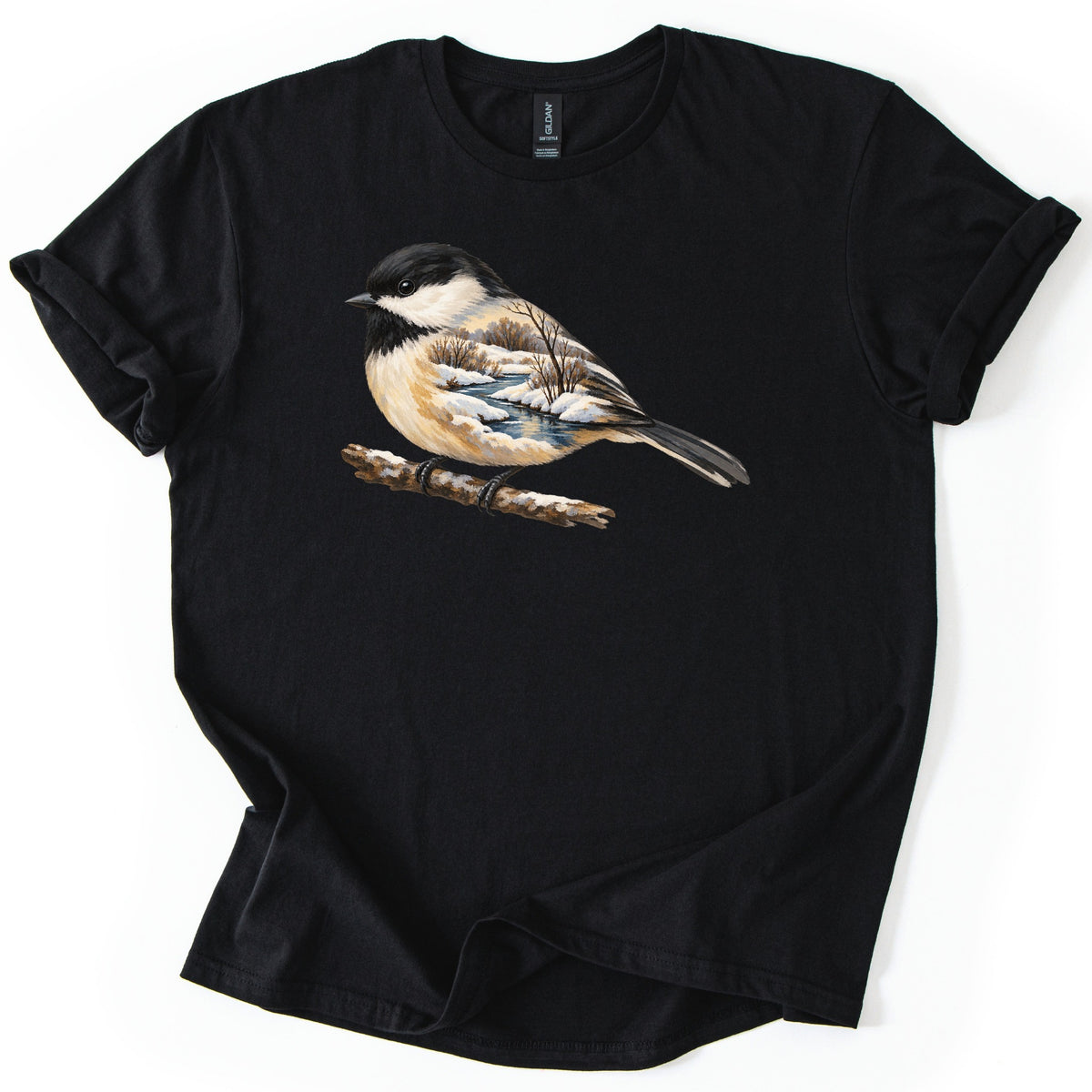 Chickadee Winter Creek Comfort Colors T-shirt in Black, laid flat. A painted black-capped chickadee perches on a snowy branch, its body filled with a winter scene of a creek, snowbanks and bare trees. No words are printed. The shirt lies on a white surface with rolled sleeves.