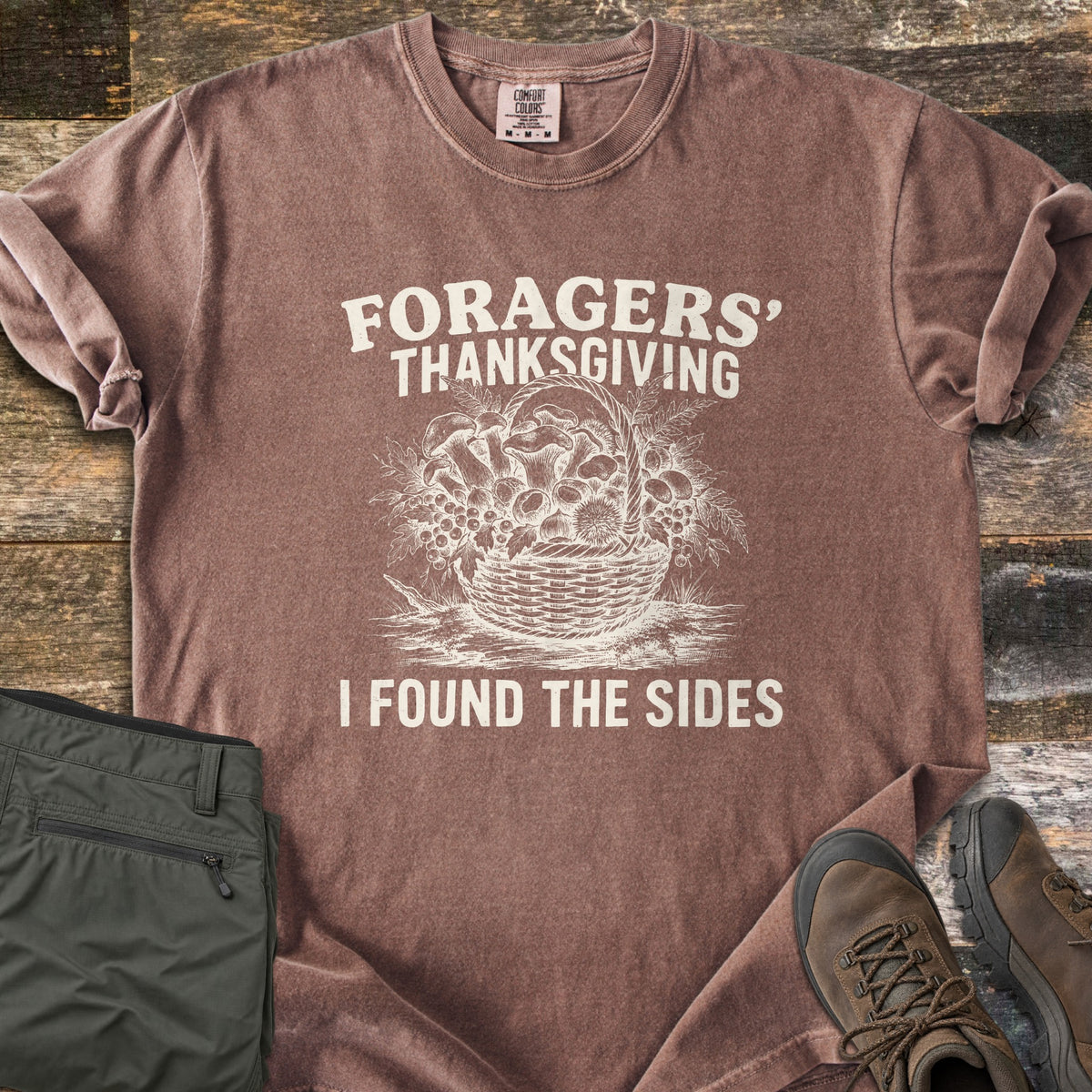 Foragers Thanksgiving Comfort Colors T-shirt in Espresso, laid flat. A cream sketch of a woven basket heaped with mushrooms, berries and ferns sits between the words Foragers' Thanksgiving and I Found the Sides. Olive hiking pants and brown boots lie beside it on rustic wood.