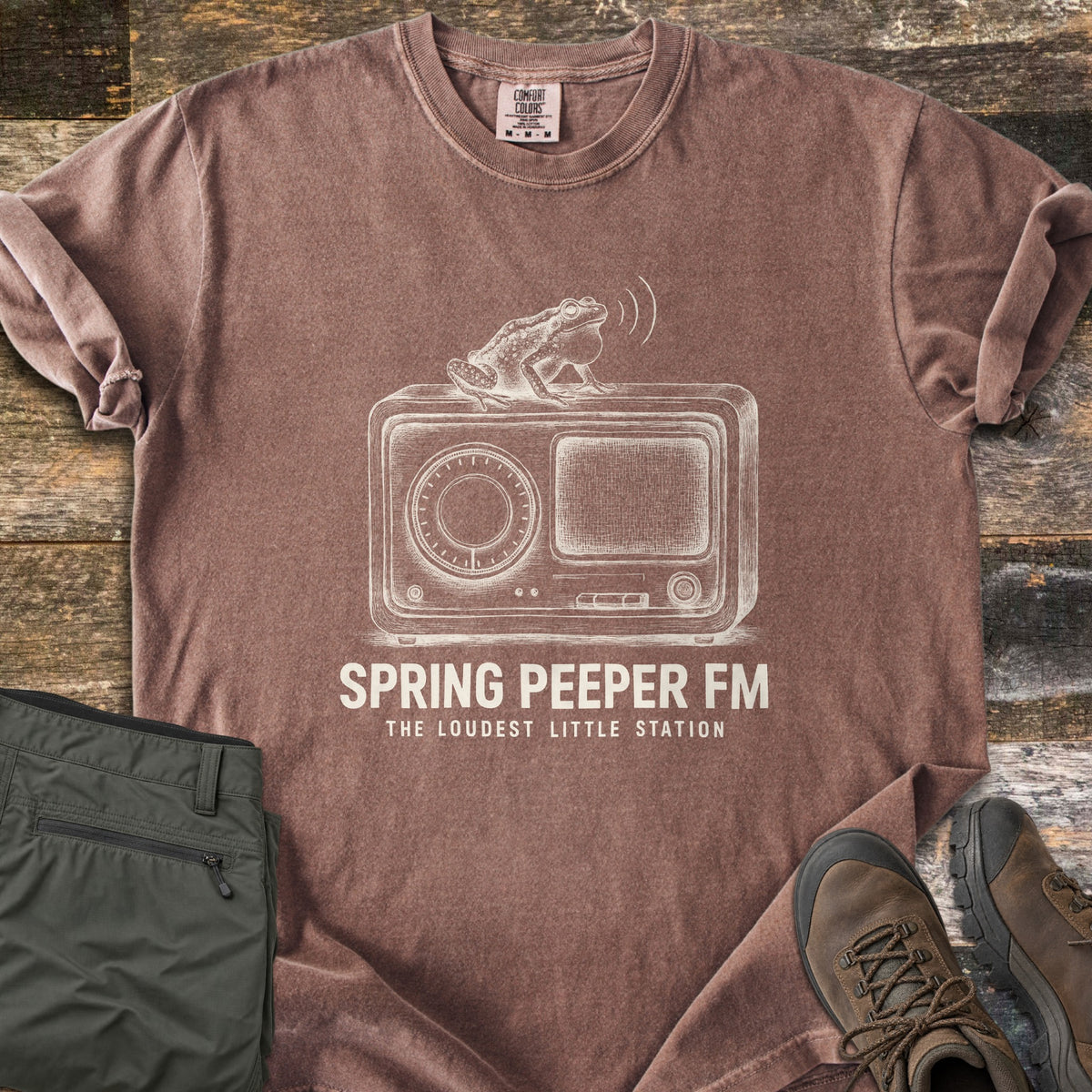 Spring Peeper FM Comfort Colors T-shirt in Espresso, laid flat. A sketch of a frog with puffed throat sits on a vintage radio, sound waves above it, over the words Spring Peeper FM, The Loudest Little Station. Sleeves are rolled on wood planks beside green pants and hiking boots.