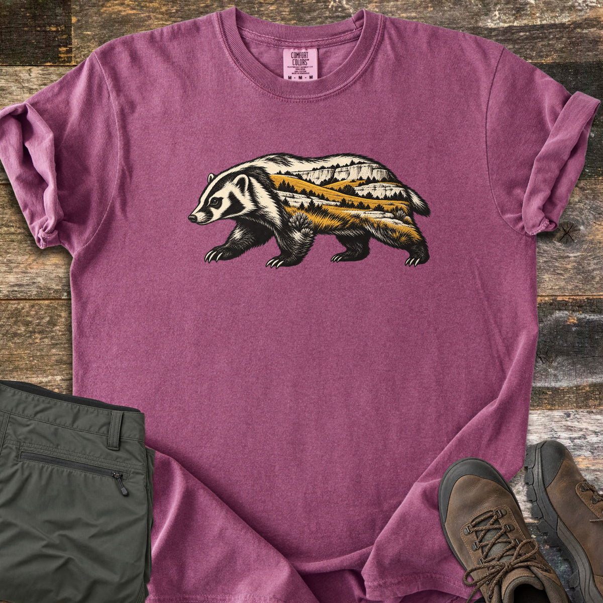 Badger Prairie Bluffs Comfort Colors T-shirt in Berry, laid flat. A walking badger silhouette filled with a prairie scene of golden grass, pine trees and white bluffs is printed on the chest. Shown on weathered wood with folded olive pants and brown hiking boots.