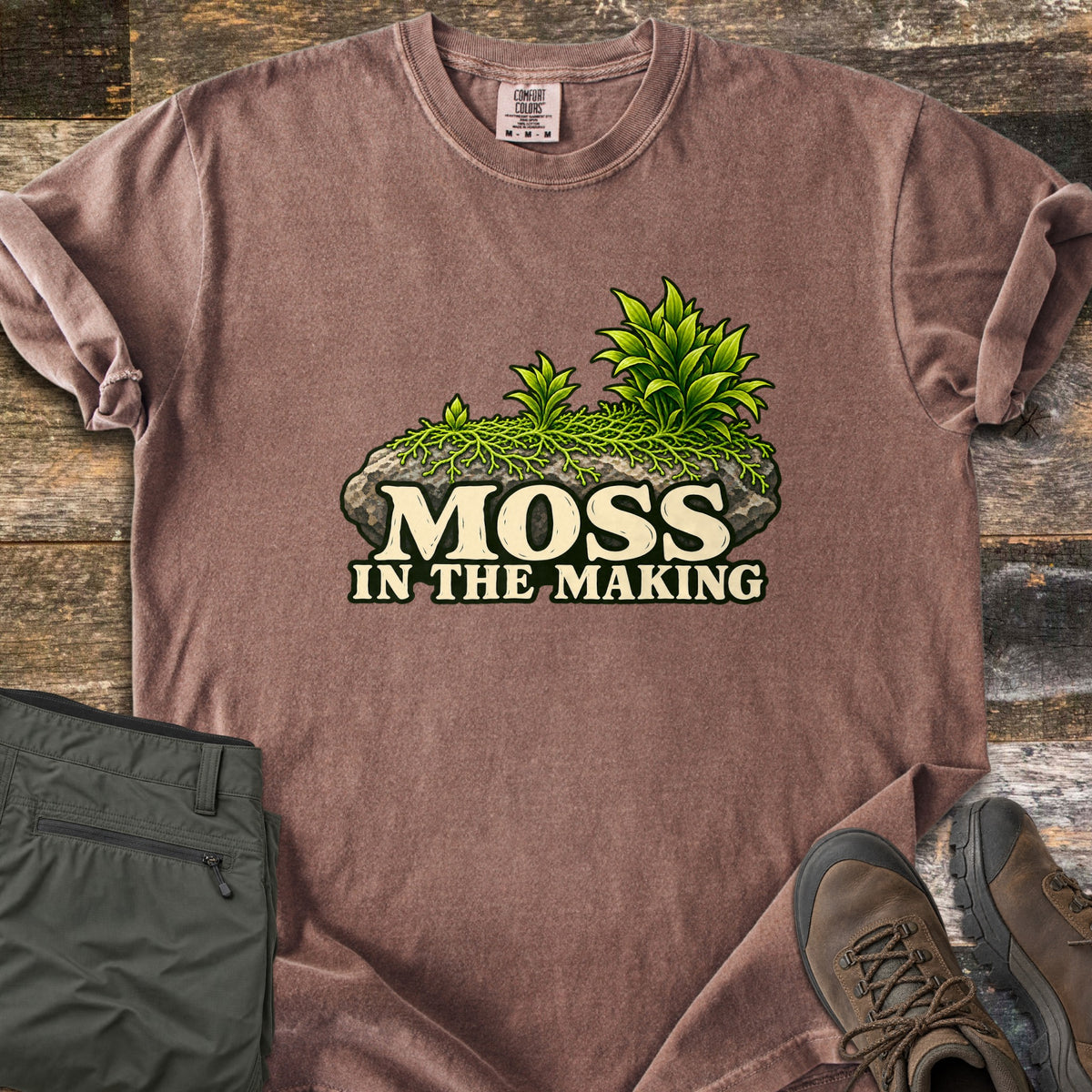 Moss in the Making Comfort Colors T-shirt in Espresso, laid flat. A mossy gray rock sprouting green leafy plants sits above the words Moss in the Making. Olive hiking pants and brown hiking boots lie beside it on weathered wood planks.