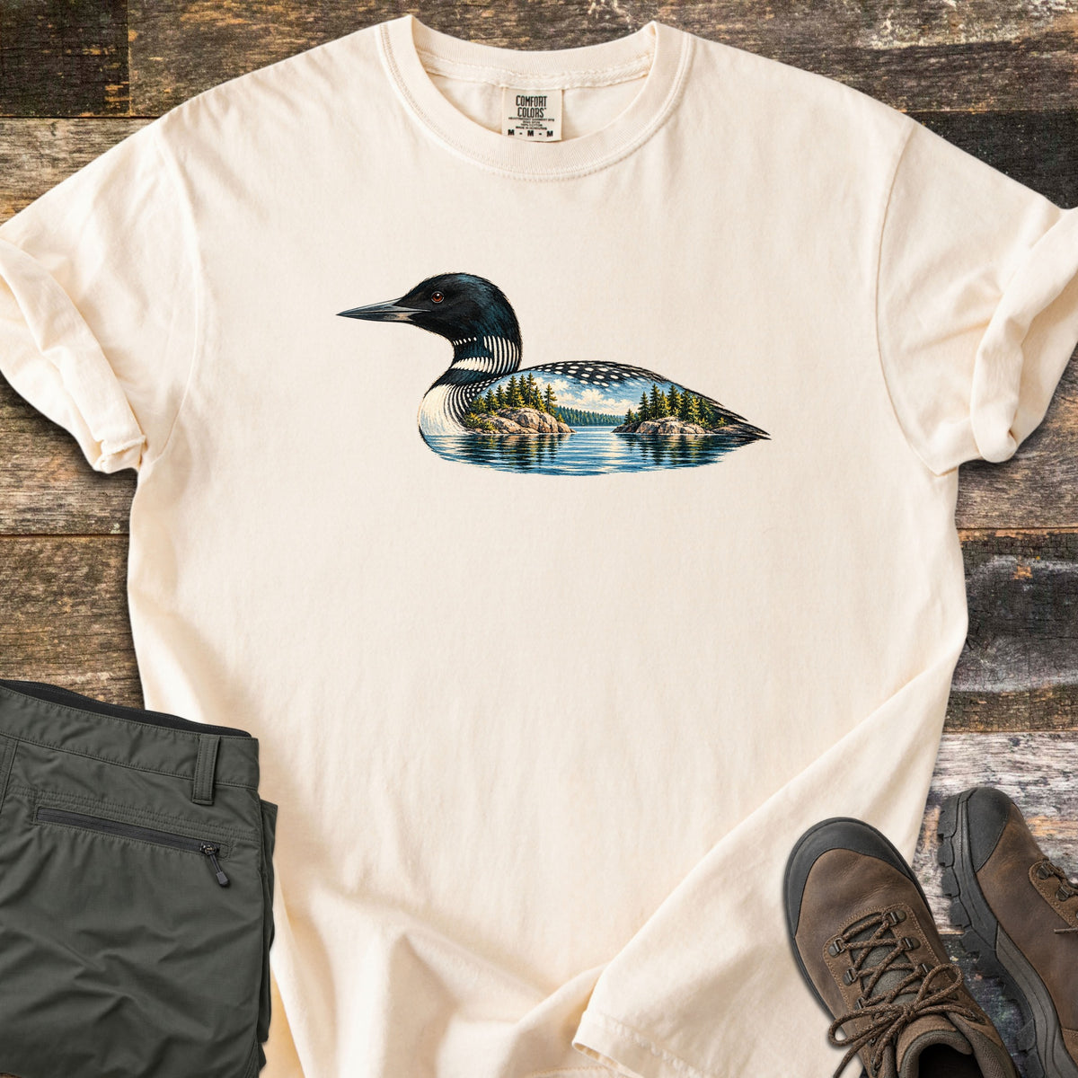 Loon Lake Comfort Colors T-shirt in Ivory, laid flat. A common loon in black and white plumage whose body holds a lake scene with pine-topped rocky islands, blue sky and reflections. Olive hiking pants and brown hiking boots sit beside it on weathered wood planks.