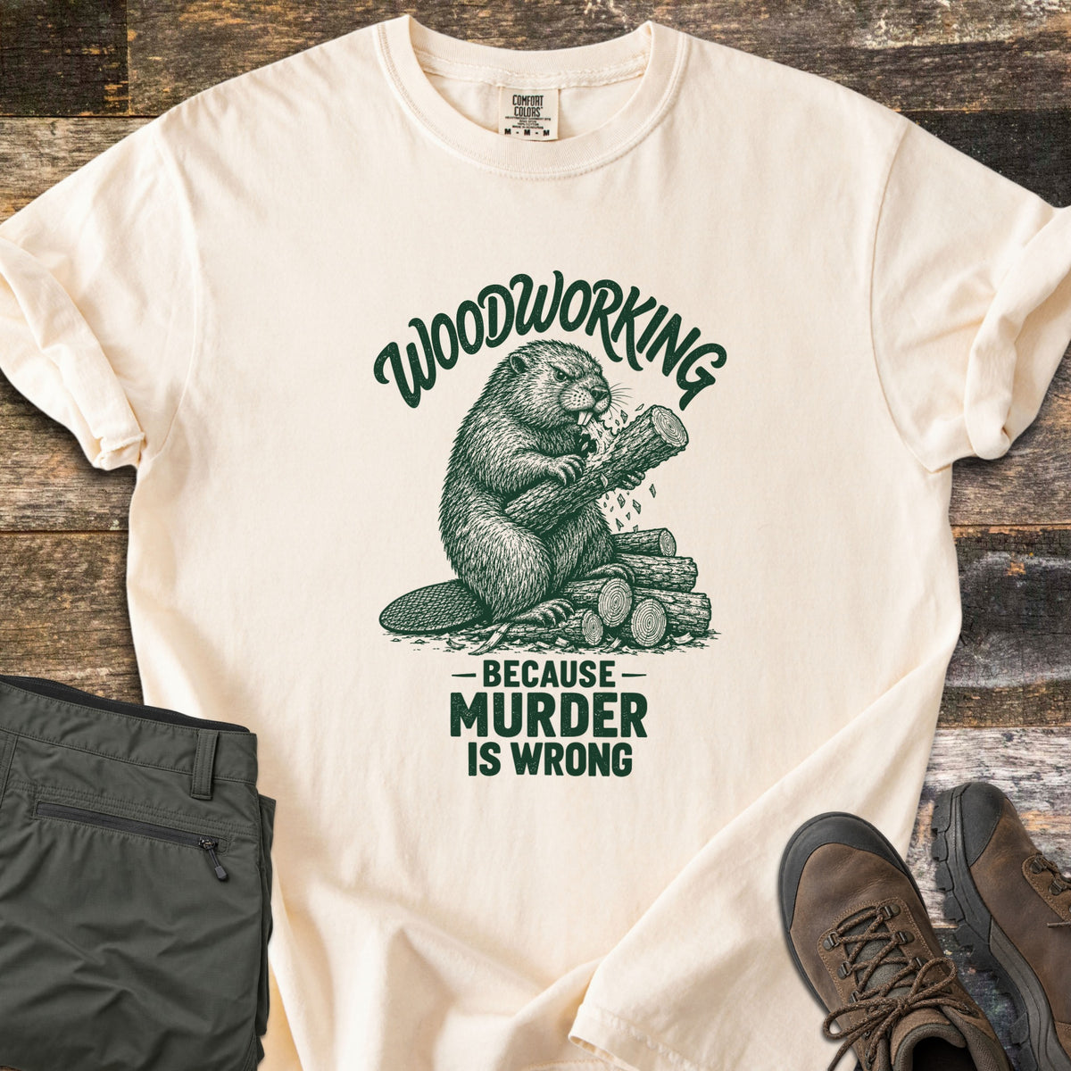 Woodworking Because Murder Is Wrong Comfort Colors T-shirt in Ivory, laid flat. A dark green sketch of a beaver gnawing a log on a woodpile, under the arched words Woodworking and above Because Murder Is Wrong. Olive cargo pants and brown hiking boots sit beside it on weathered wood planks.