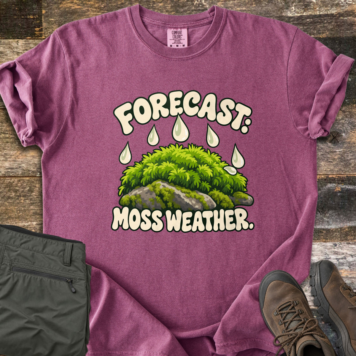 Forecast Moss Weather Comfort Colors T-shirt in Berry, laid flat. A cartoon mound of bright green moss on a gray rock, with raindrops falling from the words Forecast: above and Moss Weather. below. Olive hiking pants and brown hiking boots sit beside it on weathered wood boards.