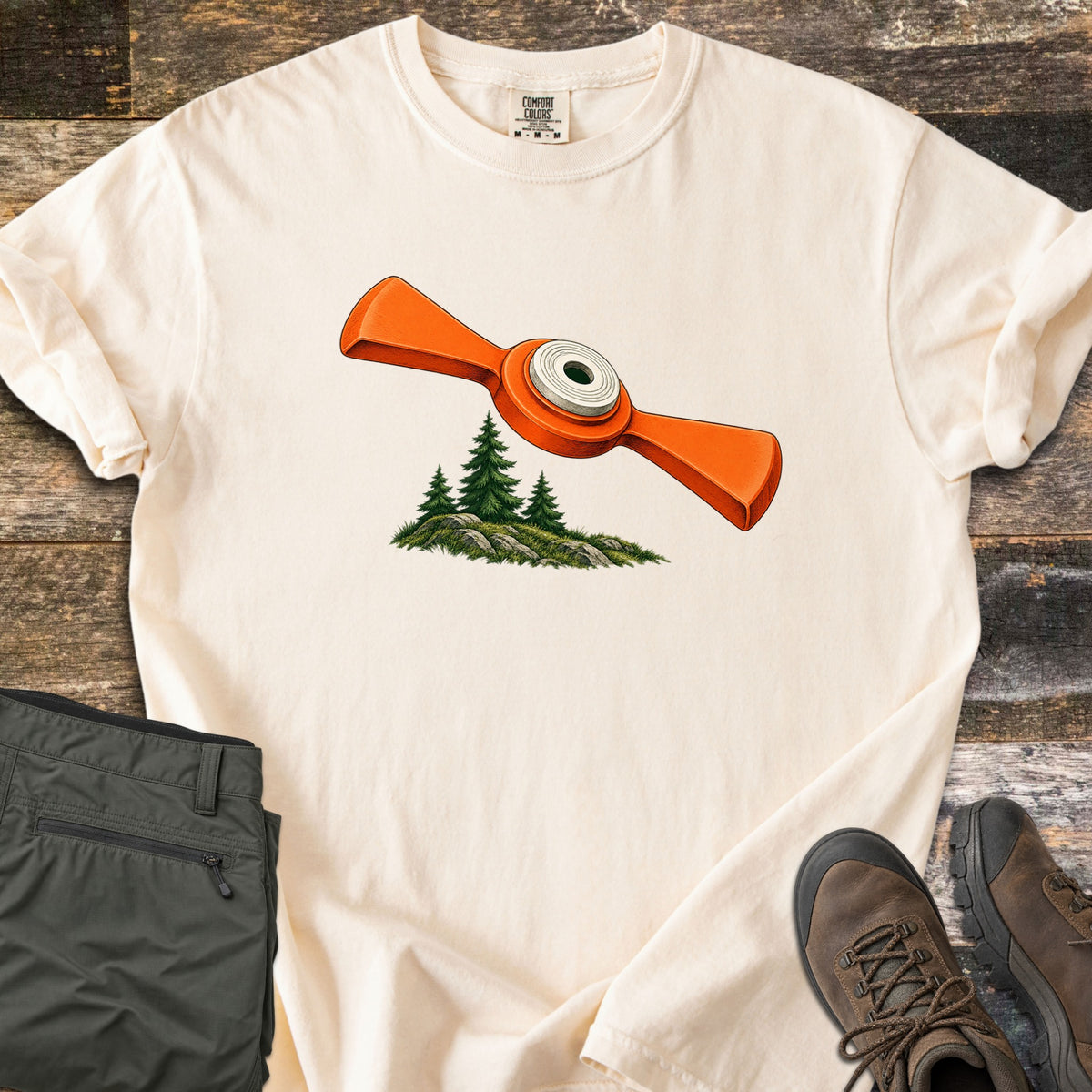 Winged Target Over the Pines Comfort Colors T-shirt in Ivory, laid flat. An orange winged clay target with a white center soars above three pine trees on a mossy, rocky knoll, with no words. Folded olive hiking pants and brown hiking boots lie beside it on weathered wood.
