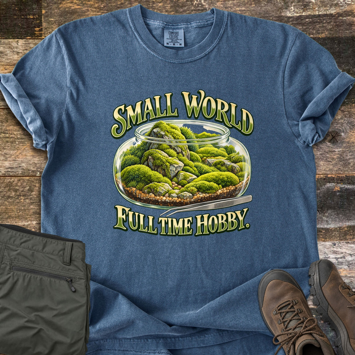 Small World Full Time Hobby Comfort Colors T-shirt in Denim, laid flat. A glass jar terrarium of green moss and rocks with tweezers sits between the words Small World and Full Time Hobby. On a wood floor beside olive cargo pants and brown hiking boots.