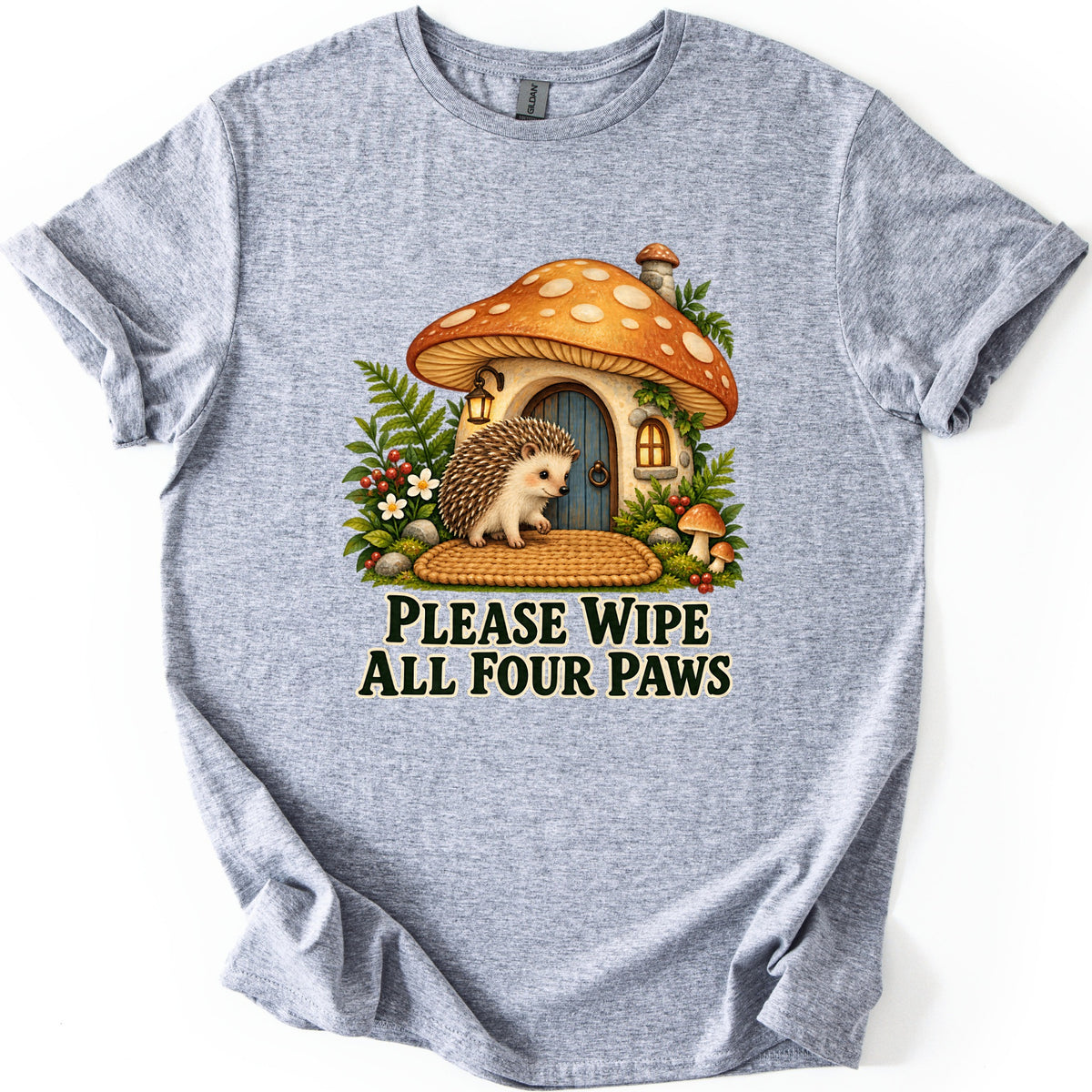 Please Wipe All Four Paws Comfort Colors T-shirt in Sport Grey, laid flat. A hedgehog on a woven doormat stands at the blue door of a mushroom cottage with an orange spotted cap, ferns, flowers and berries. Below, dark green text reads Please Wipe All Four Paws.