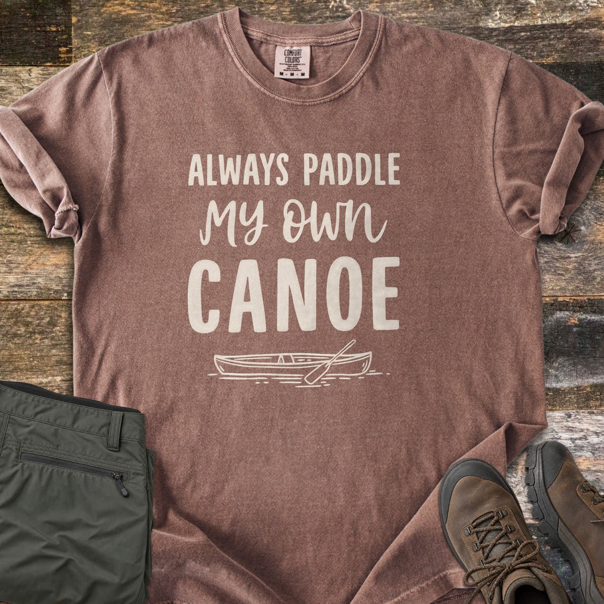 Paddle My Own Canoe Comfort Colors T-shirt in Espresso, laid flat. Cream text reads Always Paddle My Own Canoe above a line drawing of a canoe with a paddle on the water. It lies on weathered wood with rolled sleeves, folded olive hiking pants, and brown hiking boots.