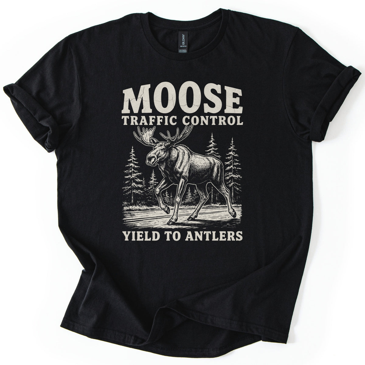 Moose Traffic Control Comfort Colors T-shirt in Black, laid flat. A cream vintage-style drawing of a bull moose crossing a road in front of pine trees, with Moose Traffic Control above and Yield to Antlers below. The sleeves are rolled, on a white background.