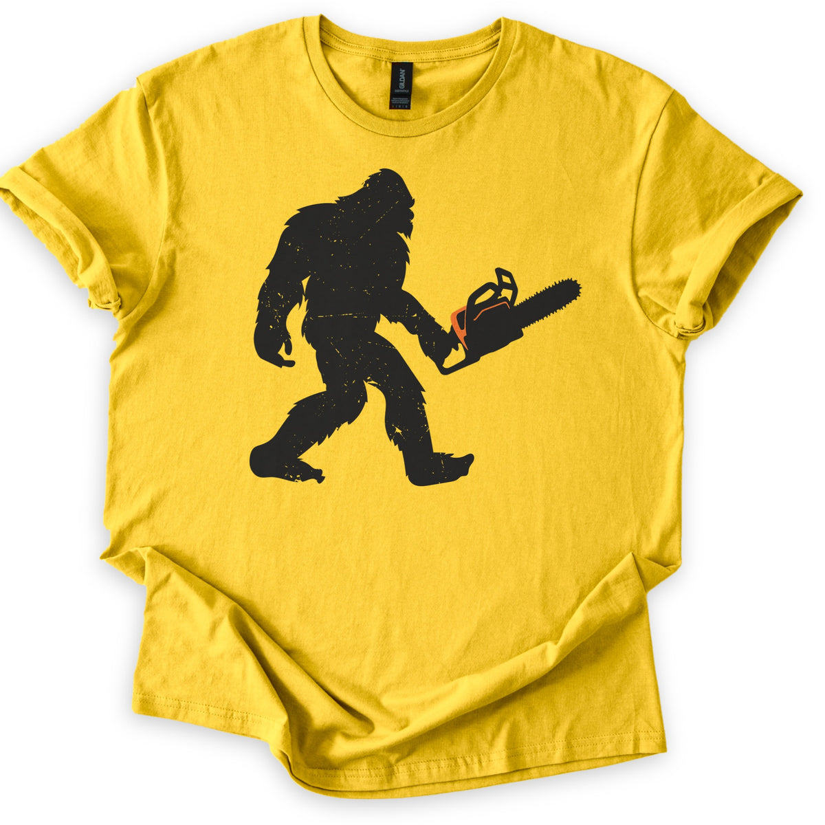 Lumber Squatch Comfort Colors T-shirt in Daisy, laid flat. A distressed black silhouette of Bigfoot walking mid-stride while holding a chainsaw with an orange accent, with no words printed. The shirt has cuffed sleeves and lies on a white background.