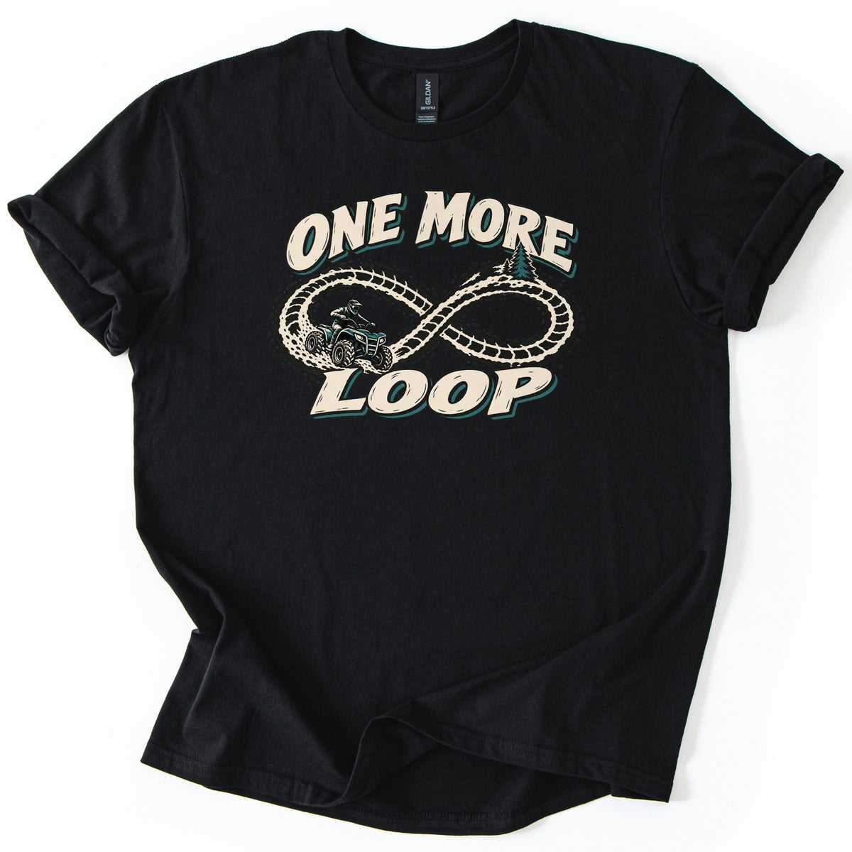 One More Loop Comfort Colors T-shirt in Black, laid flat. Cream letters reading One More Loop frame an infinity-shaped tire track with pine trees, ridden by a helmeted ATV rider. The shirt lies on a white surface with cuffed sleeves.