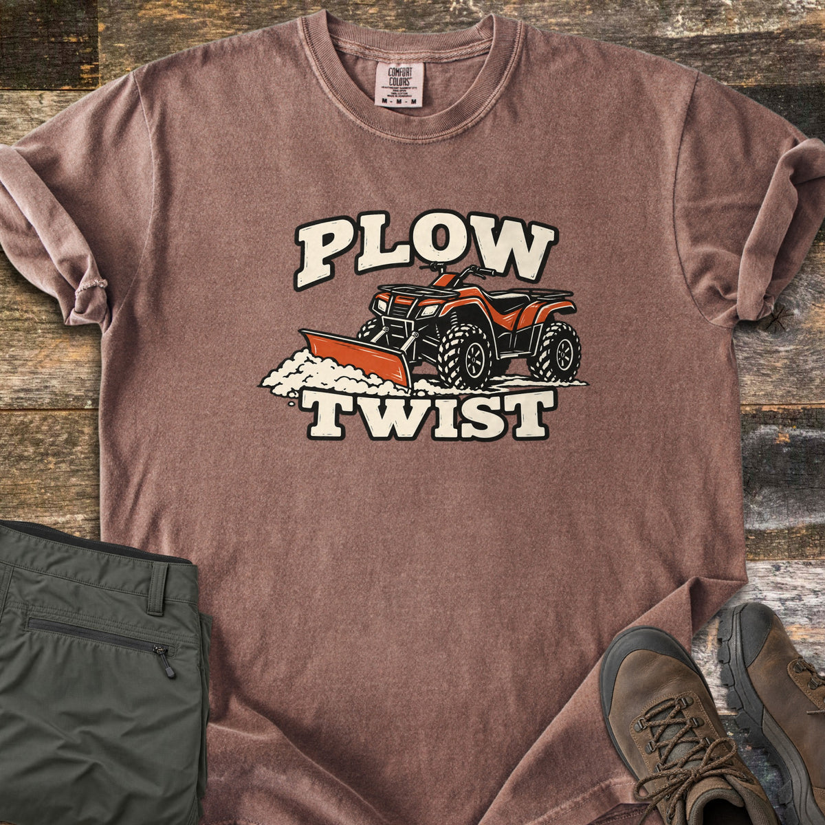 Plow Twist Comfort Colors T-shirt in Espresso, laid flat. An orange ATV with a snow plow blade pushes a pile of snow between the words Plow Twist in cream lettering. Folded olive cargo pants and brown hiking boots sit beside it on weathered wood planks.