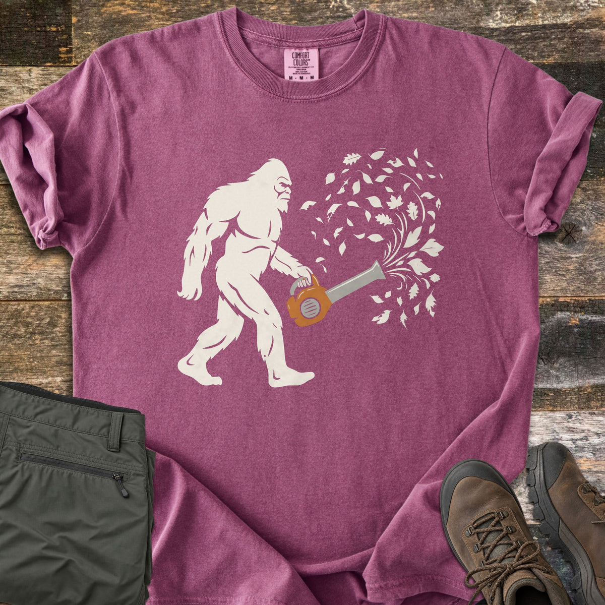 Bigfoot Leaf Blower Comfort Colors T-shirt in Berry, laid flat. A white Bigfoot silhouette walks while holding an orange leaf blower that sends a swirl of white leaves into the air. Folded olive pants and brown hiking boots sit beside it on weathered wood boards.