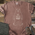 Will Hug For Vittles Comfort Colors T-shirt in Espresso, laid flat. A cream outline of a standing bear has the words Will Hug For Vittles on its belly, with two small pine trees by its feet. Folded olive hiking pants and brown hiking boots sit beside it on weathered wood.