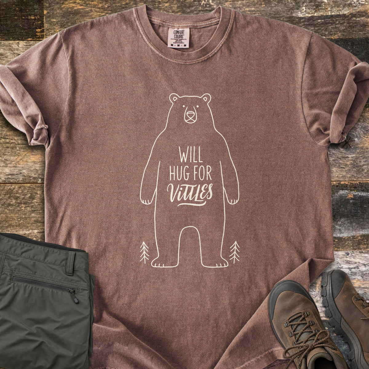 Will Hug For Vittles Comfort Colors T-shirt in Espresso, laid flat. A cream outline of a standing bear has the words Will Hug For Vittles on its belly, with two small pine trees by its feet. Folded olive hiking pants and brown hiking boots sit beside it on weathered wood.