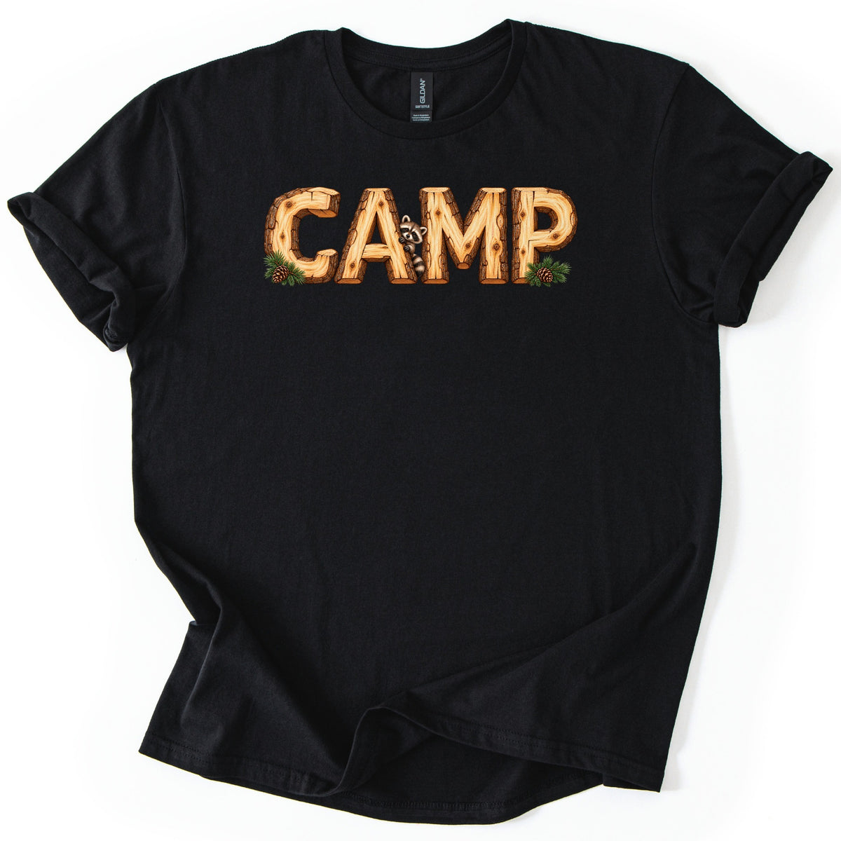 Camp Comfort Colors T-shirt in Black, laid flat. The word CAMP is printed in log letters with bark edges and wood grain, a raccoon peeking out beside the A, and pine sprigs with pinecones at each end. The shirt lies on a white surface with cuffed sleeves.