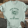 You're Welcome For The Sunrise Comfort Colors T-shirt in Bay, laid flat. A green etched rooster stands on a wooden post crowing, with a rising sun behind it, under the curved words You're Welcome and above For The Sunrise. Folded olive pants and brown hiking boots sit on weathered wood.