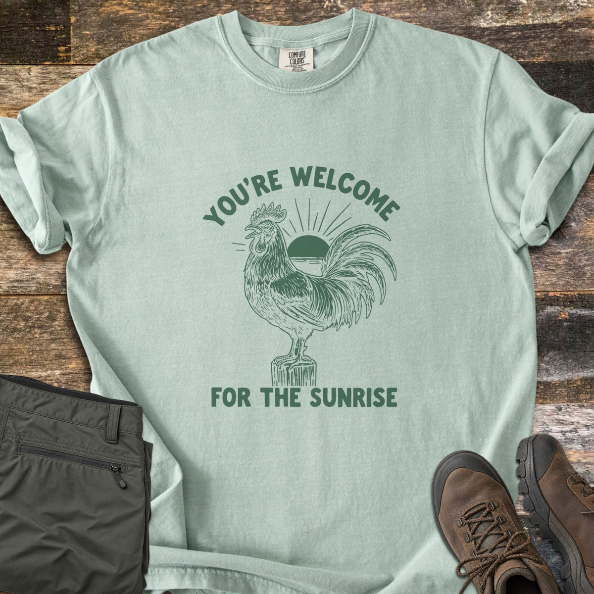 You're Welcome For The Sunrise Comfort Colors T-shirt in Bay, laid flat. A green etched rooster stands on a wooden post crowing, with a rising sun behind it, under the curved words You're Welcome and above For The Sunrise. Folded olive pants and brown hiking boots sit on weathered wood.