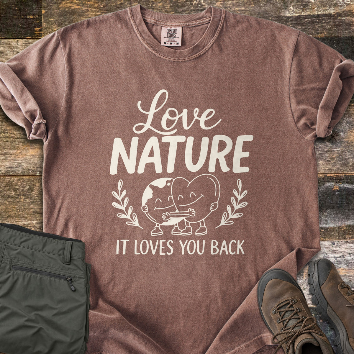 Nature Loves You Back Comfort Colors T-shirt in Espresso, laid flat. Cream print reads Love Nature above a smiling cartoon Earth and heart hugging between leafy sprigs, with It Loves You Back below. It lies on weathered wood beside olive hiking pants and brown boots.
