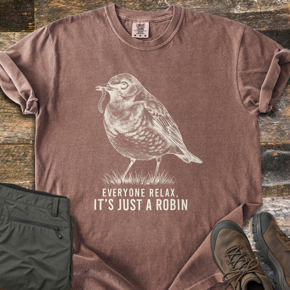 Everyone Relax It's Just A Robin Comfort Colors T-shirt in Espresso, laid flat. A sketched robin with a worm in its beak stands on grass above the words Everyone Relax, It's Just A Robin, in cream ink. Olive cargo pants and brown hiking boots lie beside it on weathered wood.