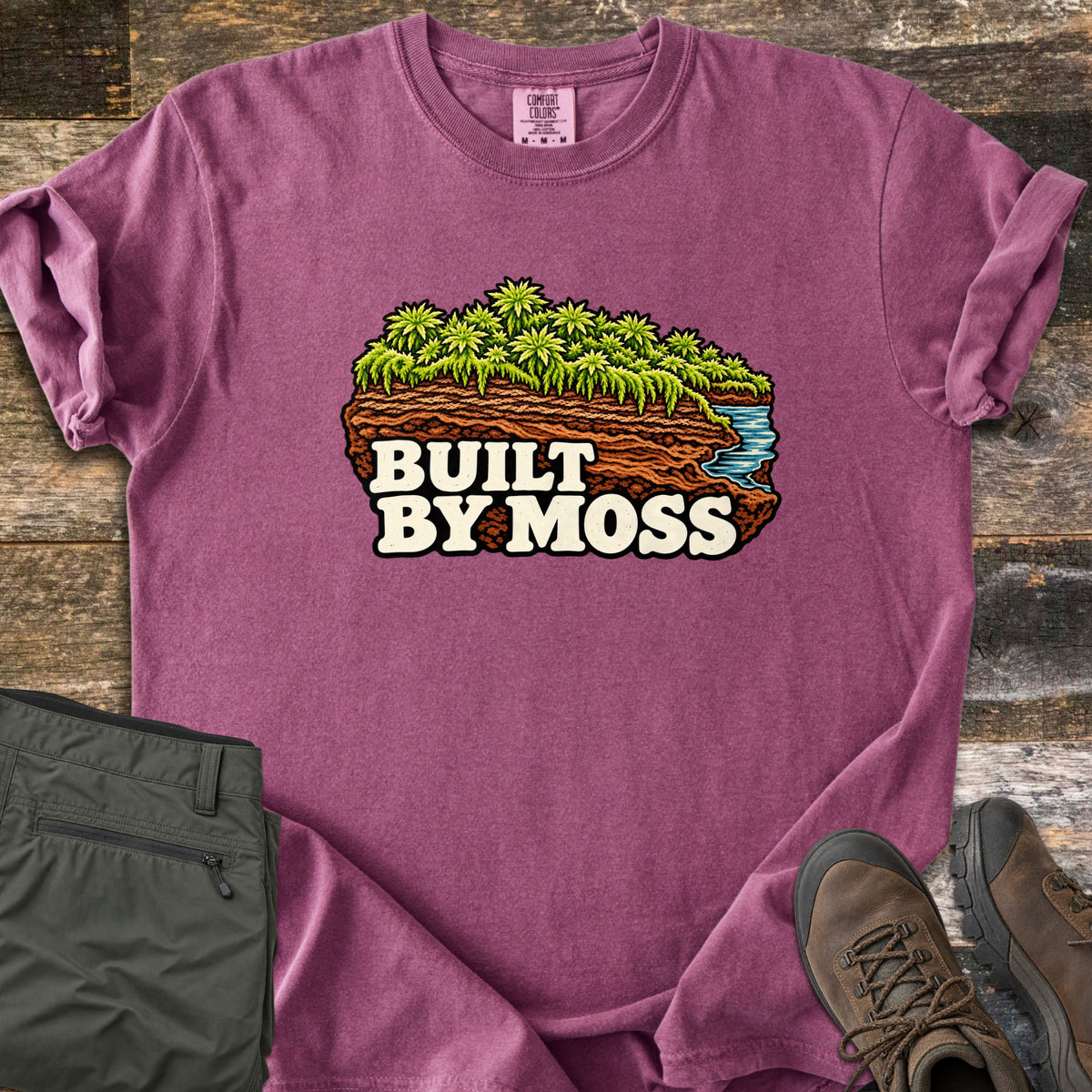 Built by Moss Comfort Colors T-shirt in Berry, laid flat. An illustrated chunk of brown soil topped with bright green moss and a small blue stream, with the words Built by Moss. Shown on weathered wood with folded olive hiking pants and brown hiking boots.