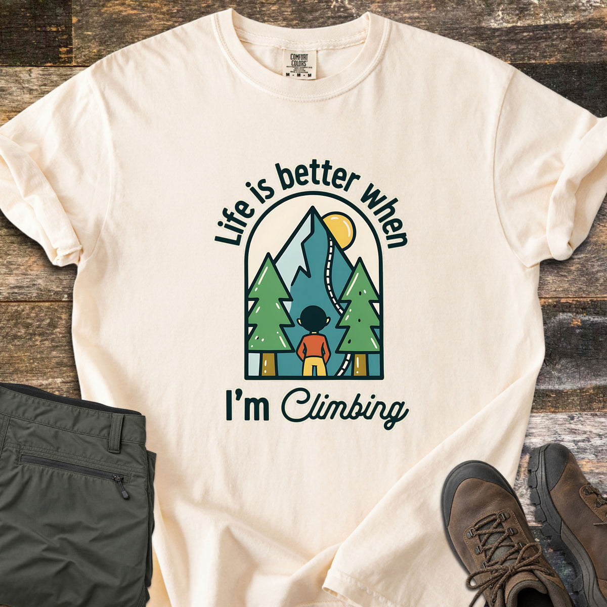 Better When I'm Climbing Comfort Colors T-shirt in Ivory, laid flat. An arched scene of a person facing a mountain with a winding trail, pine trees and a yellow sun, with the words Life is better when I'm Climbing. Gray pants and brown hiking boots lie on a wood floor.