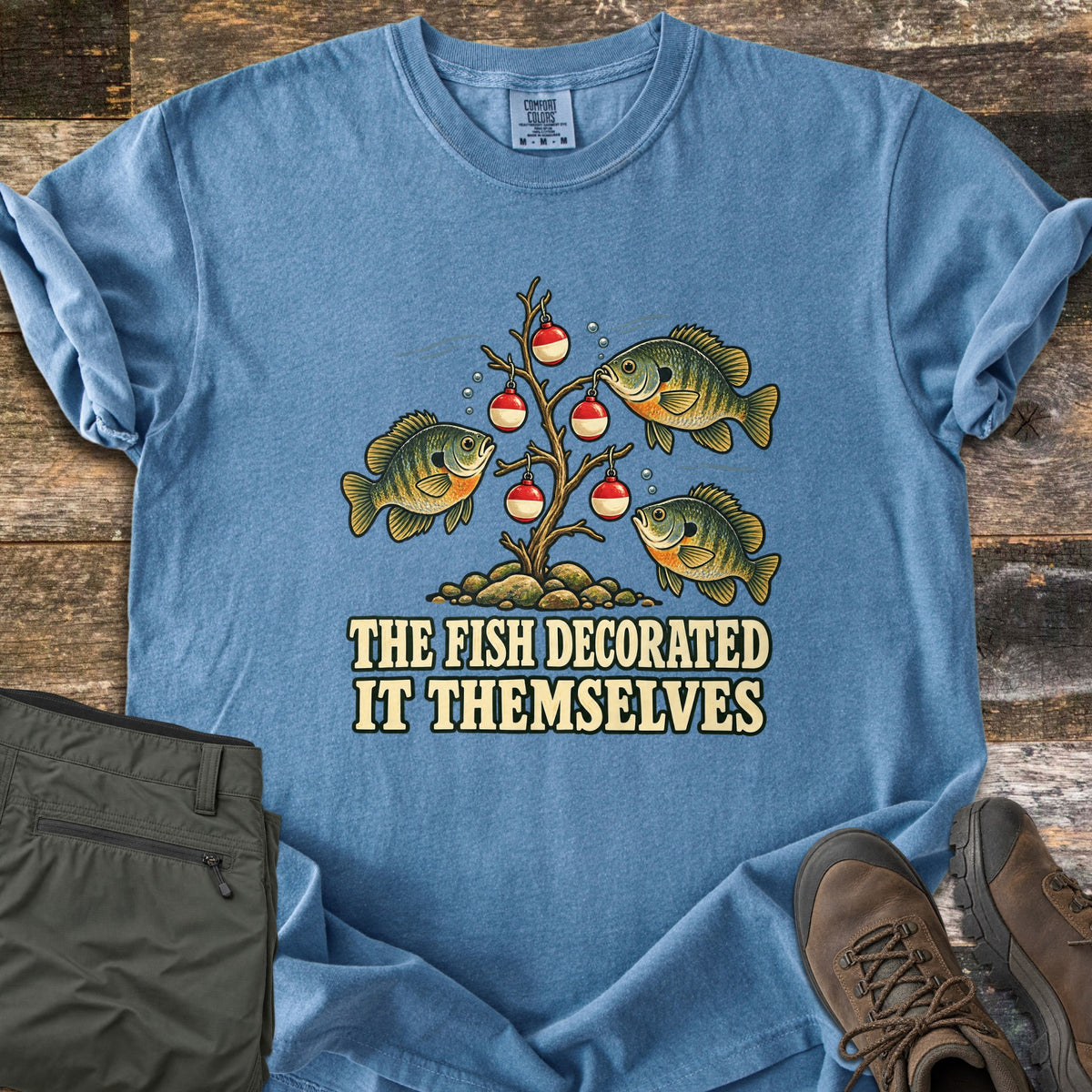 The Fish Decorated It Themselves Comfort Colors T-shirt in Blue Jean, laid flat. Three bluegill fish hang red and white fishing bobbers like ornaments on a bare underwater branch above the words The Fish Decorated It Themselves. Olive pants and brown hiking boots sit on weathered wood.