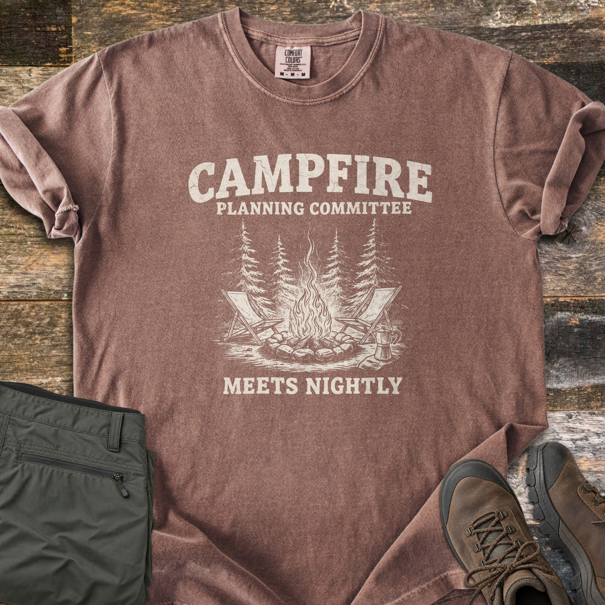 Campfire Planning Committee Comfort Colors T-shirt in Espresso, laid flat. Cream print reads Campfire Planning Committee above a sketch of a stone-ringed campfire with two folding chairs, pine trees and a coffee pot, then Meets Nightly. Olive pants and brown hiking boots lie on weathered wood.