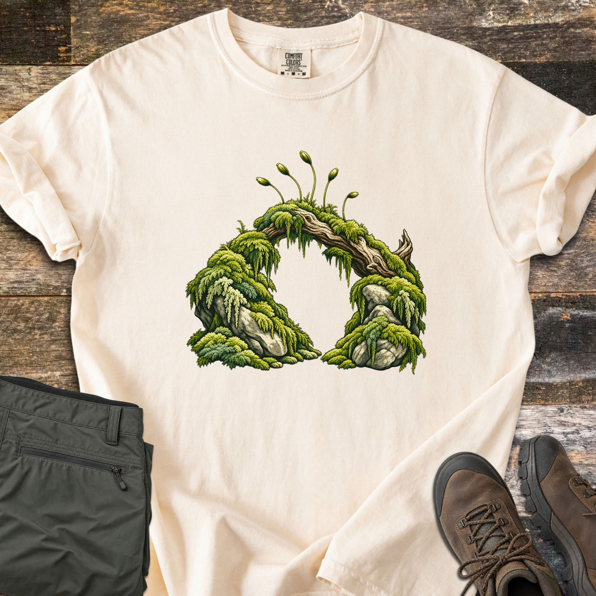 Moss Arch Comfort Colors T-shirt in Ivory, laid flat. An illustrated arch of moss-covered rocks joined by a weathered branch, with slender moss sporophytes sprouting on top; no words. Sleeves are rolled, on a wood background with olive pants and brown hiking boots.