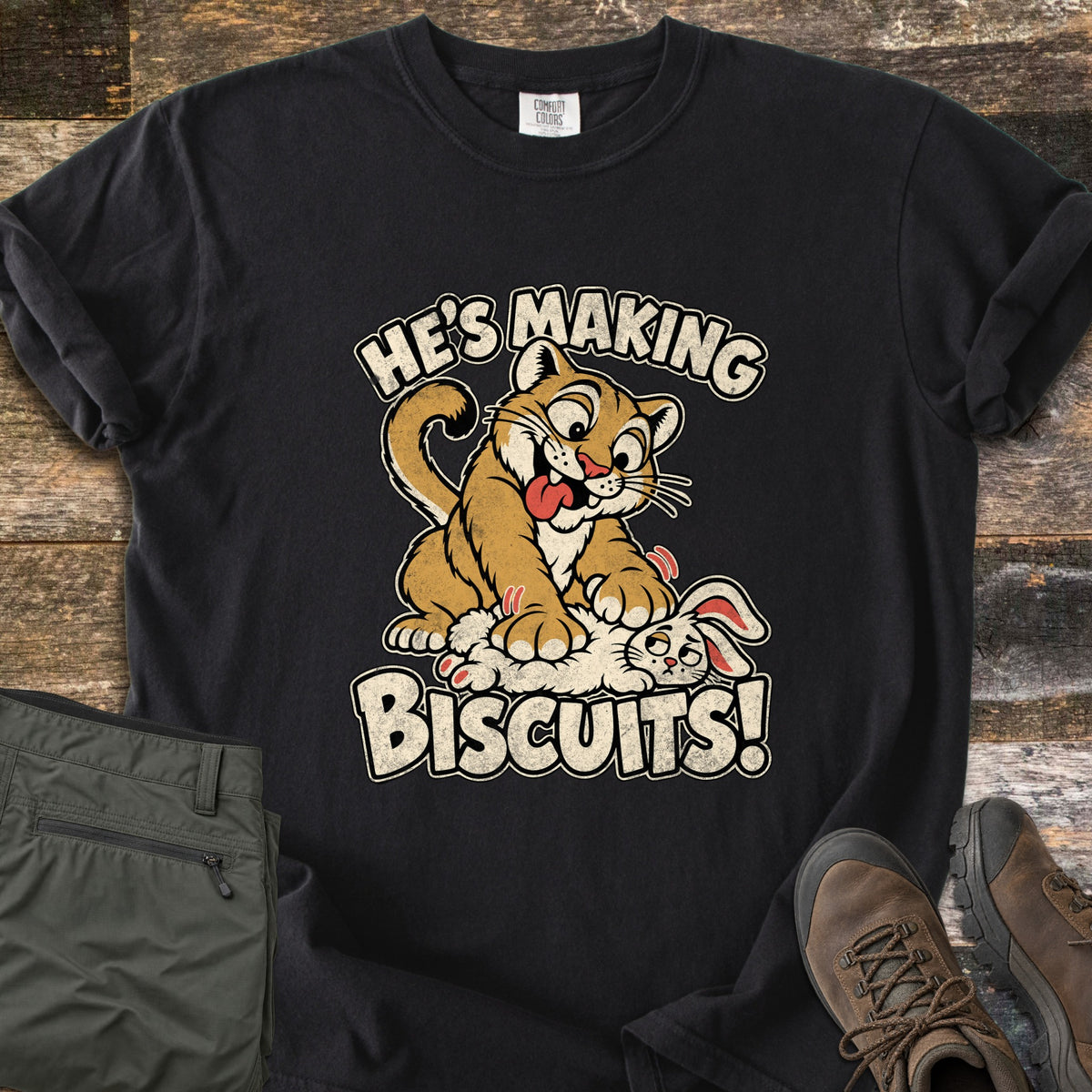 He's Making Biscuits Comfort Colors T-shirt in Black, laid flat. A cartoon cougar with its tongue out kneads a dazed white rabbit with its paws, between the words He's Making Biscuits! Olive pants and brown hiking boots sit beside it on weathered wood planks.