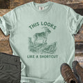 This Looks Like A Shortcut Comfort Colors T-shirt in Bay, laid flat. A dark green etching of a horned goat standing on a cliff ledge over a canyon, with the words This Looks arched above and Like A Shortcut below. Olive hiking pants and brown hiking boots lie beside it on dark wood.